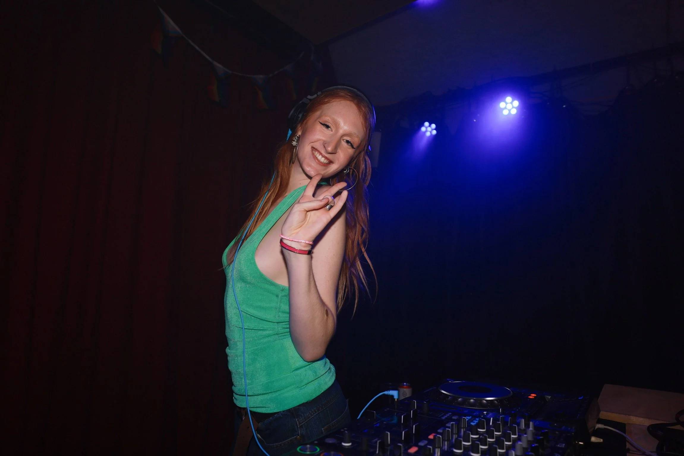 A young woman with red hair, wearing a green tank top and headphones, smiles while DJing in a dark room with blue stage lights.