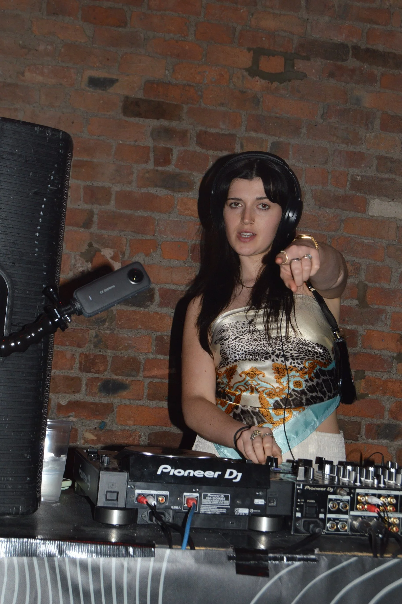 A female DJ with black hair and multiple rings on her fingers, wearing headphones, is pointing towards the camera behind a Pioneer DJ controller in a brick-walled setting.