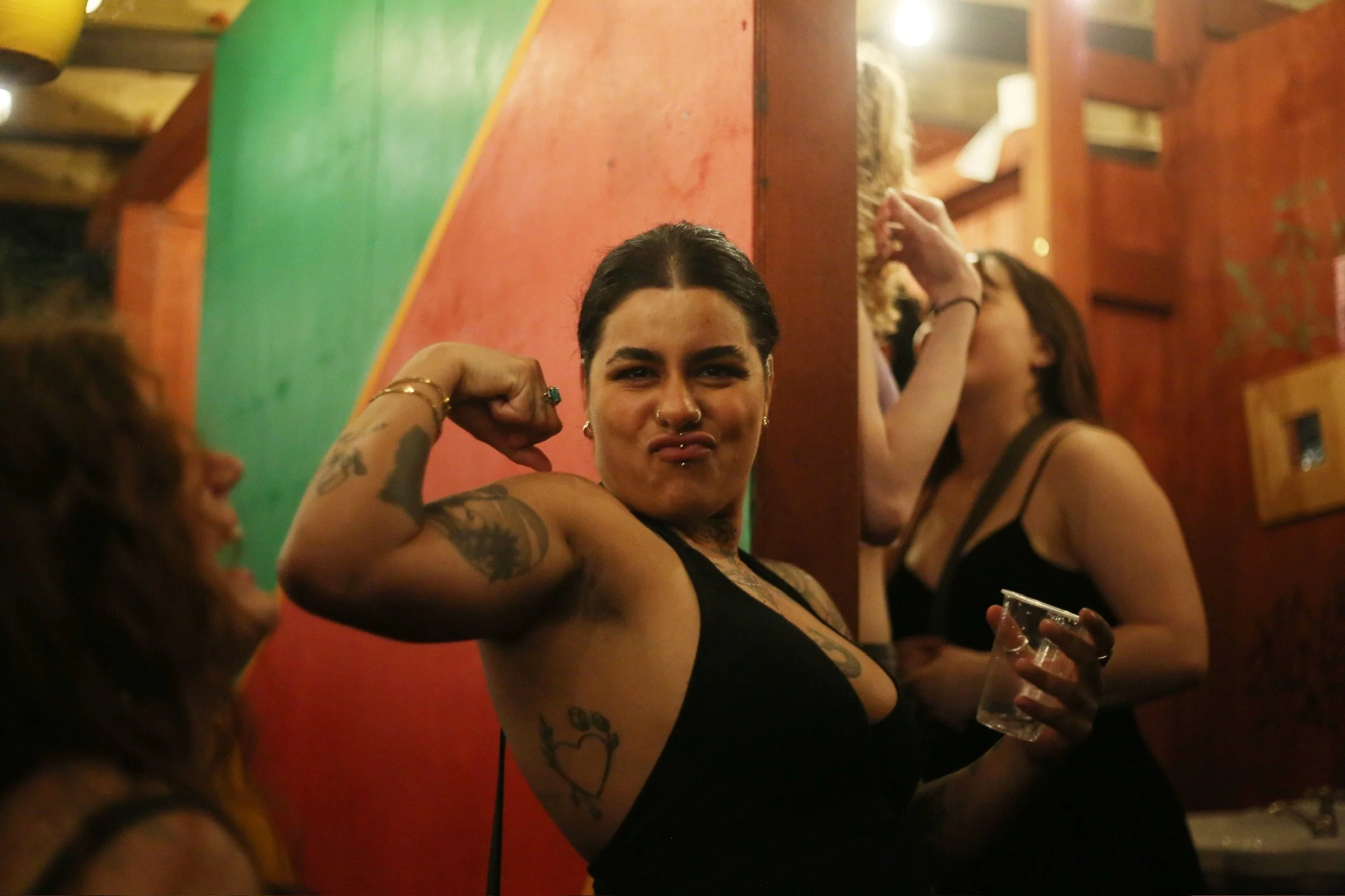 A woman with dark hair and tattoos flexing her arm in a social setting, holding a drink, with other people in the background.