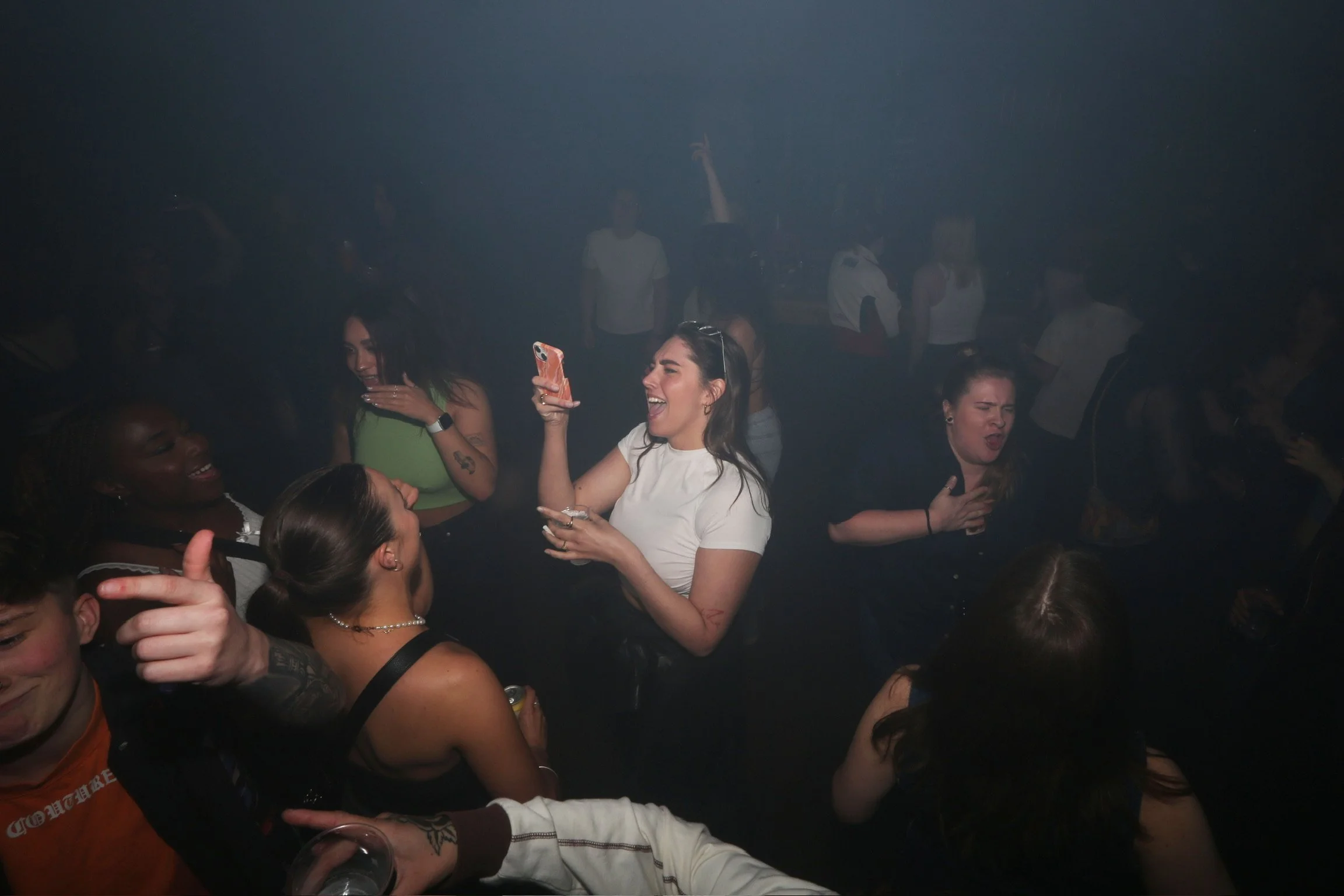 Group of people dancing and enjoying themselves at a party or nightclub, with some holding drinks and one person taking a selfie.