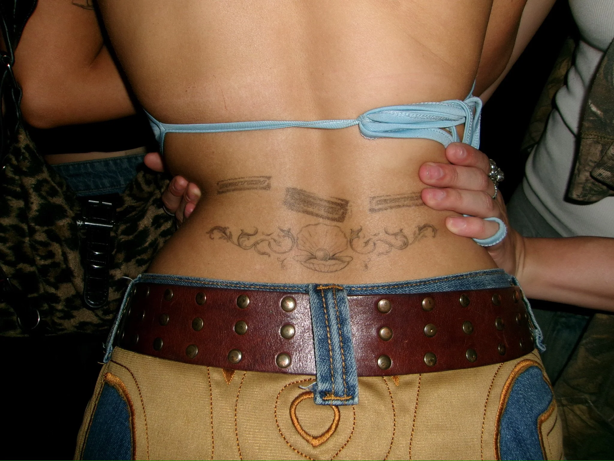 Close-up of a person's torso and waist, wearing a blue string bikini top and decorated jeans with embroidery. The person has tattoos visible on their lower abdomen and is holding their waist. A leopard print jacket and jewelry are partially visible o