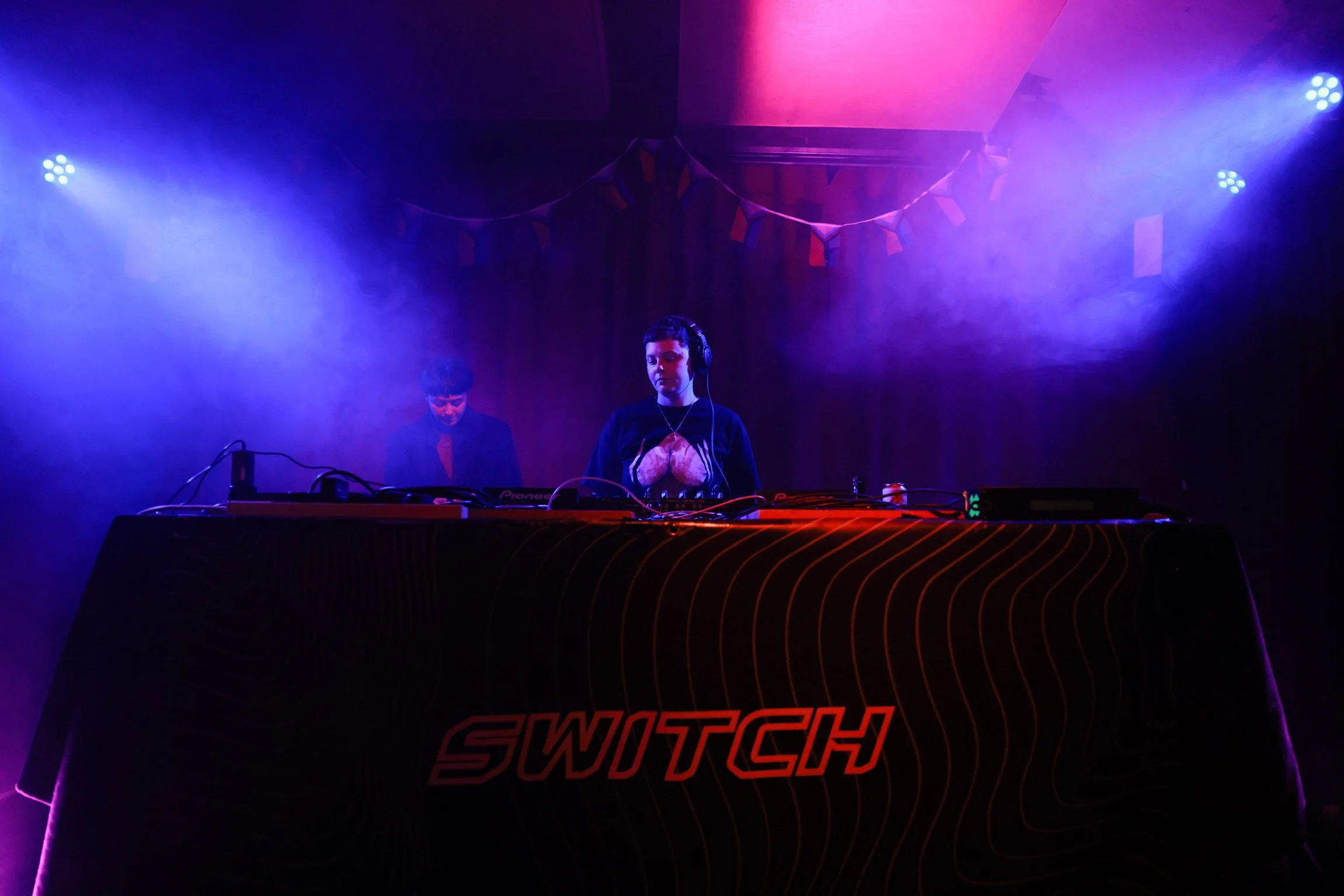 Two DJ performers are on stage under purple and pink lighting, with one wearing headphones and standing behind a Pioneer DJ controller, and the other looking down at the equipment. The stage is decorated with banners and fog, with a large Swatch logo
