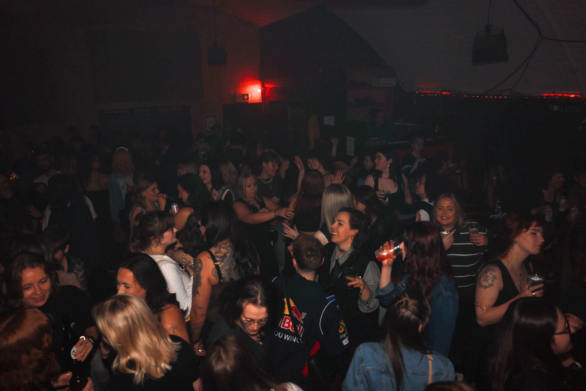 Crowd of people at a nighttime party or concert, some holding drinks, with dark lighting and a red exit sign visible in the background.