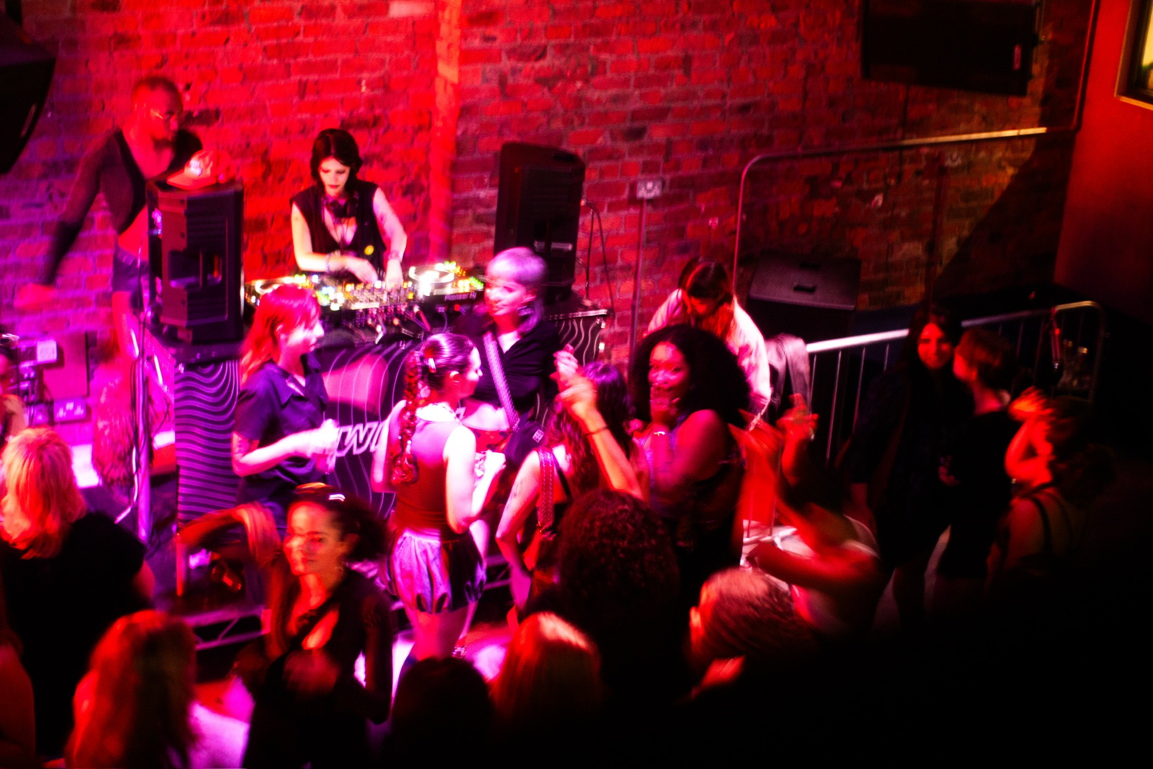 Crowd of people dancing and socializing at a nightclub with red brick walls and purple lighting, with a DJ playing music in the background.