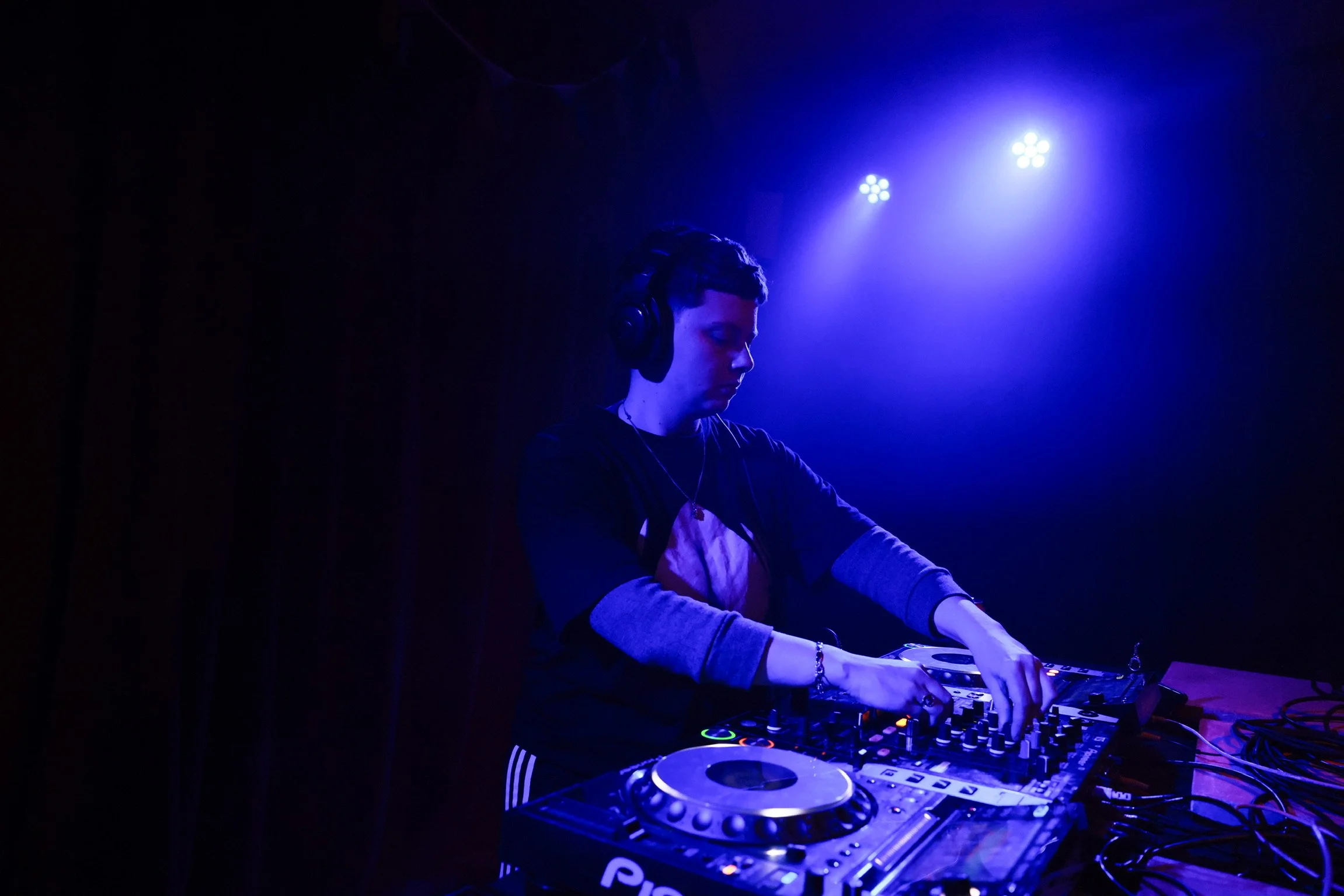A young male DJ wearing headphones performs in a dark room with blue and purple lighting, using professional DJ equipment.