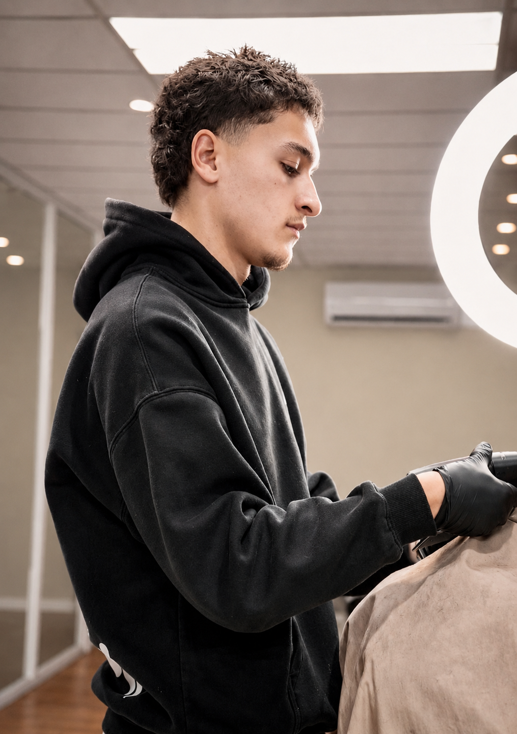 A young man with curly brown hair wearing a black hoodie and black gloves, focused on a task, inside a modern room with a large illuminated mirror.