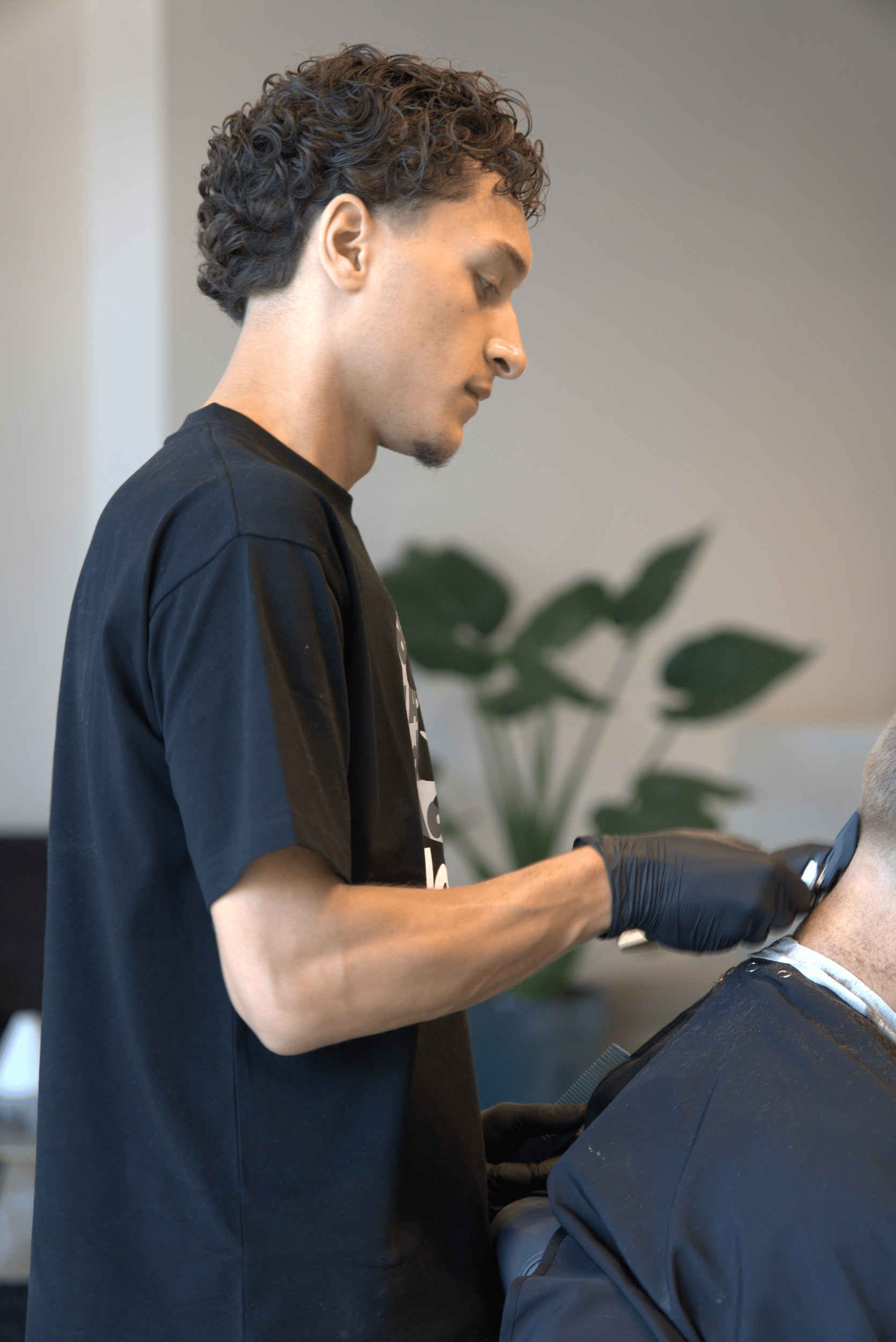 A young man with curly hair and tan skin wearing a black T-shirt and black gloves, working at a salon or barber shop, possibly giving a haircut or styling a customer.