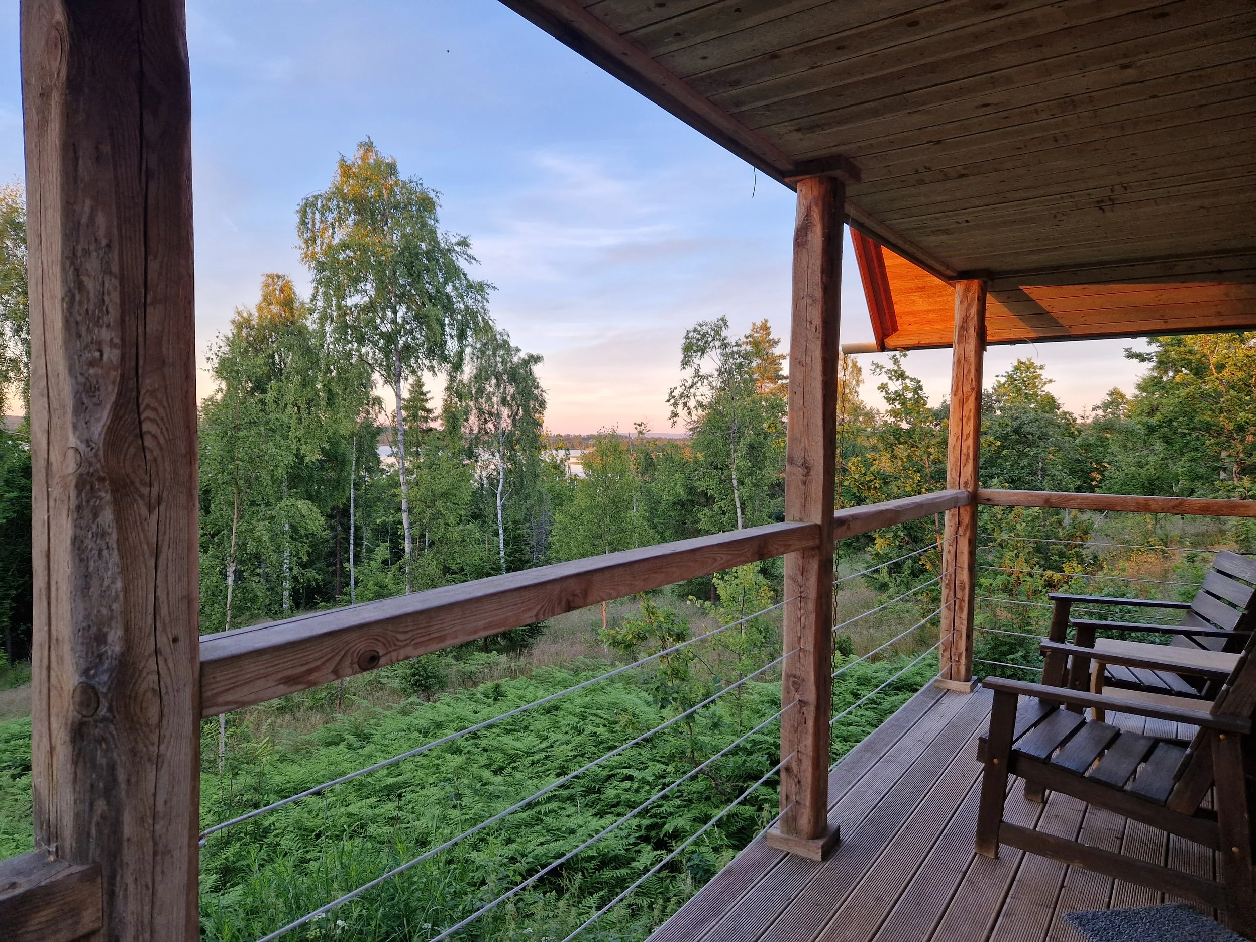 Utsikt från en träaltan med ett staket av trä och metallkabel, omgiven av gröna träd och ett lugnt vattendrag i bakgrunden, under en klar kvällssol.
