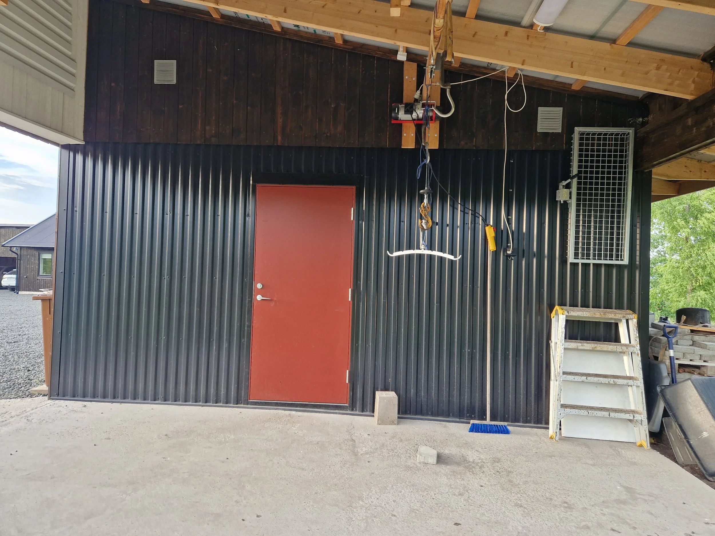 En byggarbetsplats med en svart och röd dörr, verktyg och byggmaterial runt omkring. Trä- och metallstrukturer synliga, gröna träd i bakgrunden.
