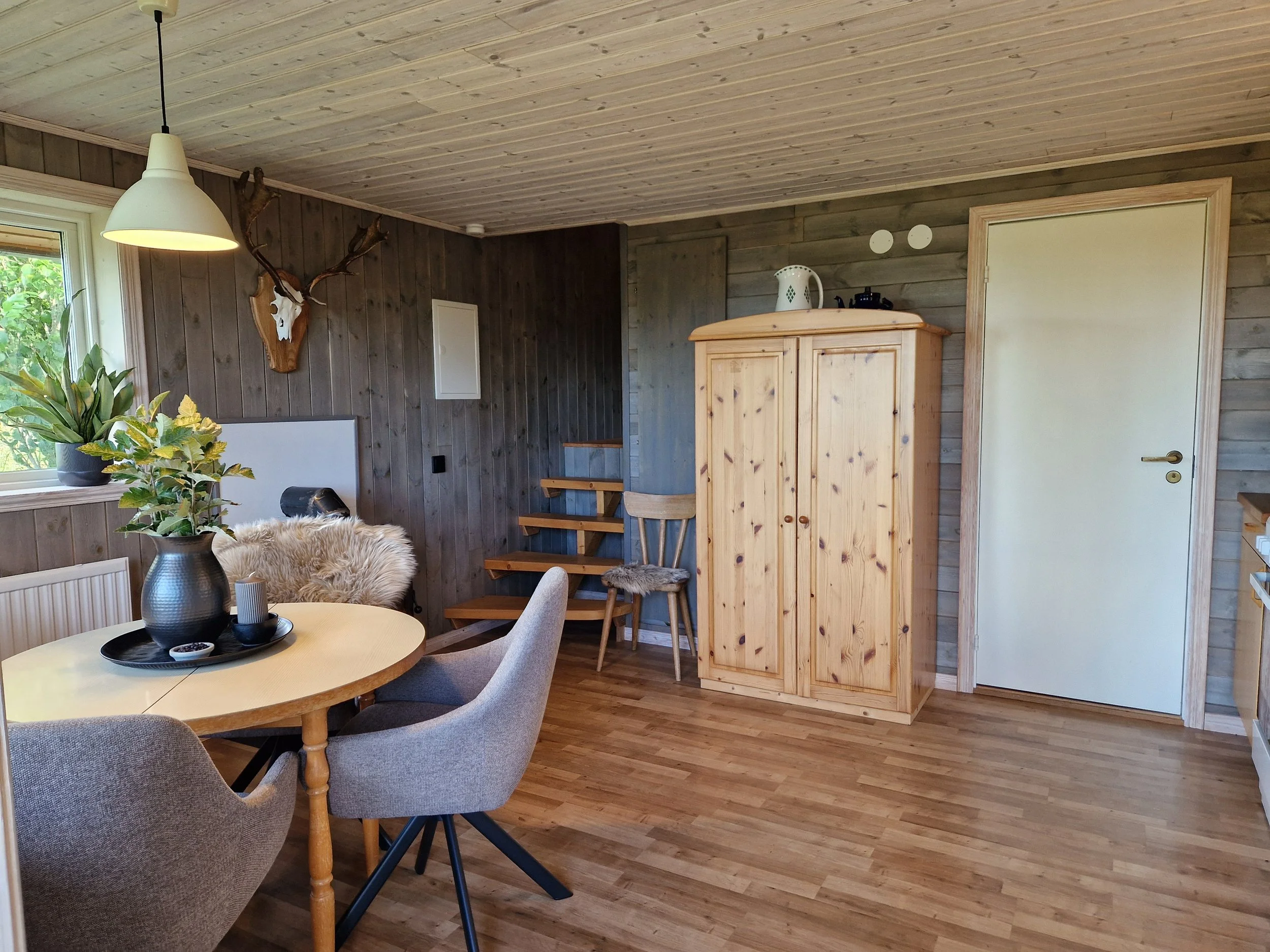 Ett rustikt kök med träväggar och tak, ett runt matbord med tre stolar, en växt i svart kruka, en träskåp, samt en vit vägglampa och en dörr.