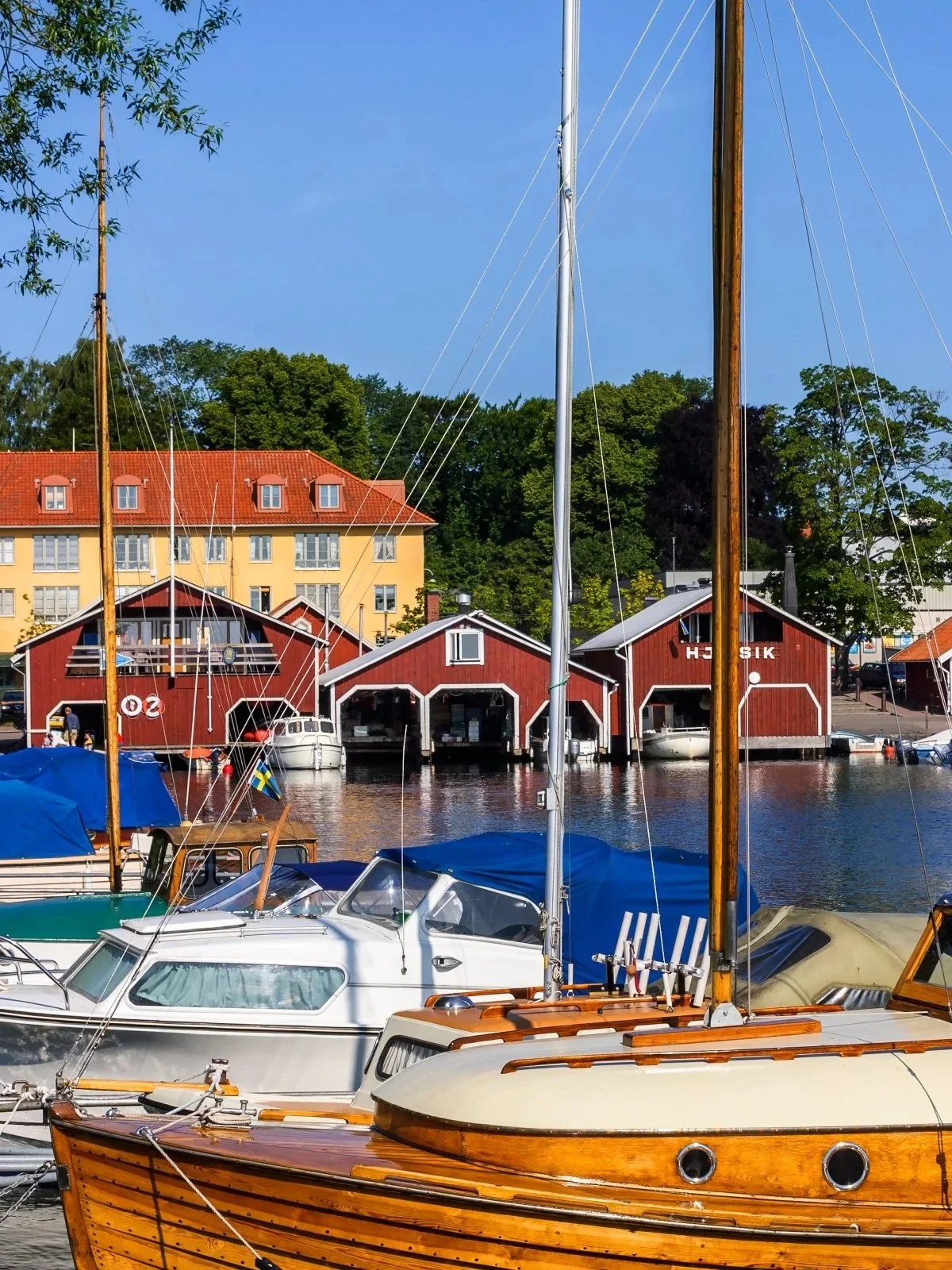 Båtar som är förtöjda vid en marina med röda båthus i bakgrunden, en gul byggnad och gröna träd, under en klar blå himmel.