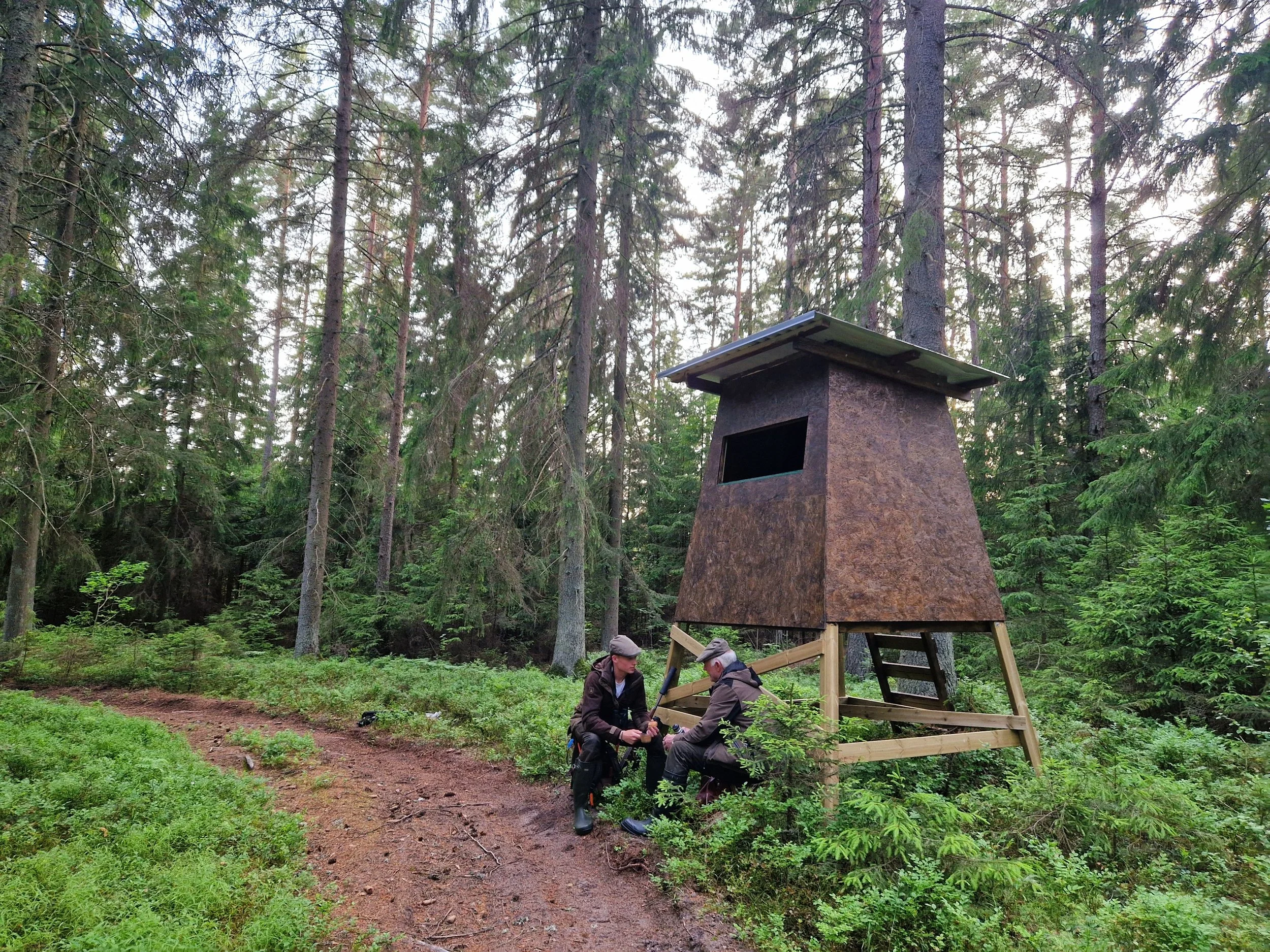 Två personer som arbetar tillsammans vid en träställning i en skog, med en rökugn eller grill.