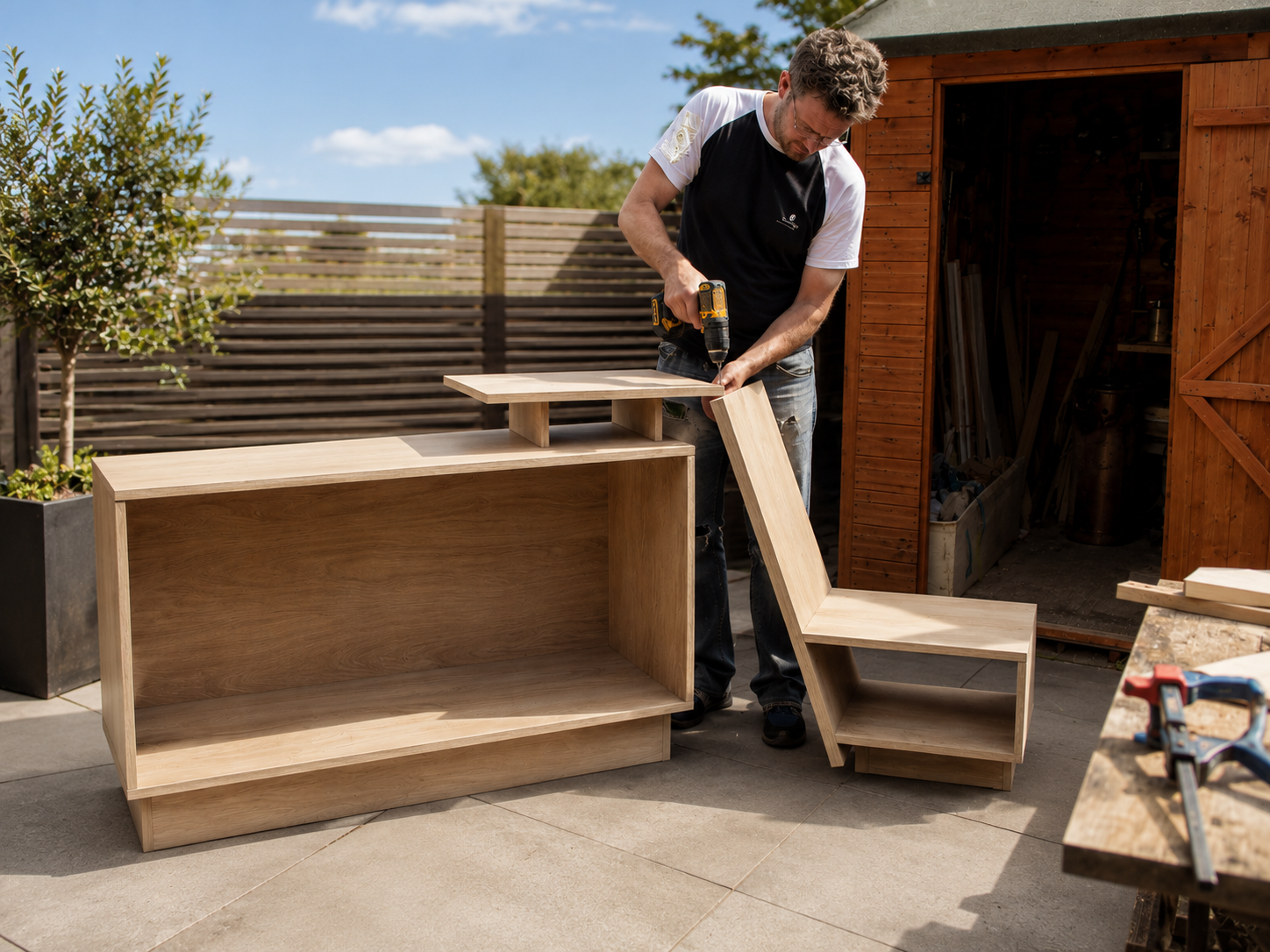 A man is assembling wooden furniture outdoors, using a cordless drill on a piece of wood, with a partially completed wooden shelf storage units showing project development.