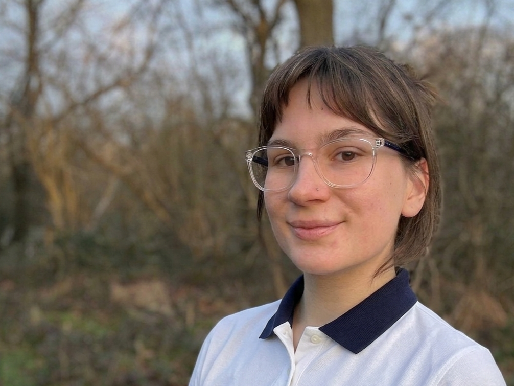 A young woman with short brown hair, wearing clear glasses and a white collared shirt with navy accents, standing outdoors with trees in the background.
