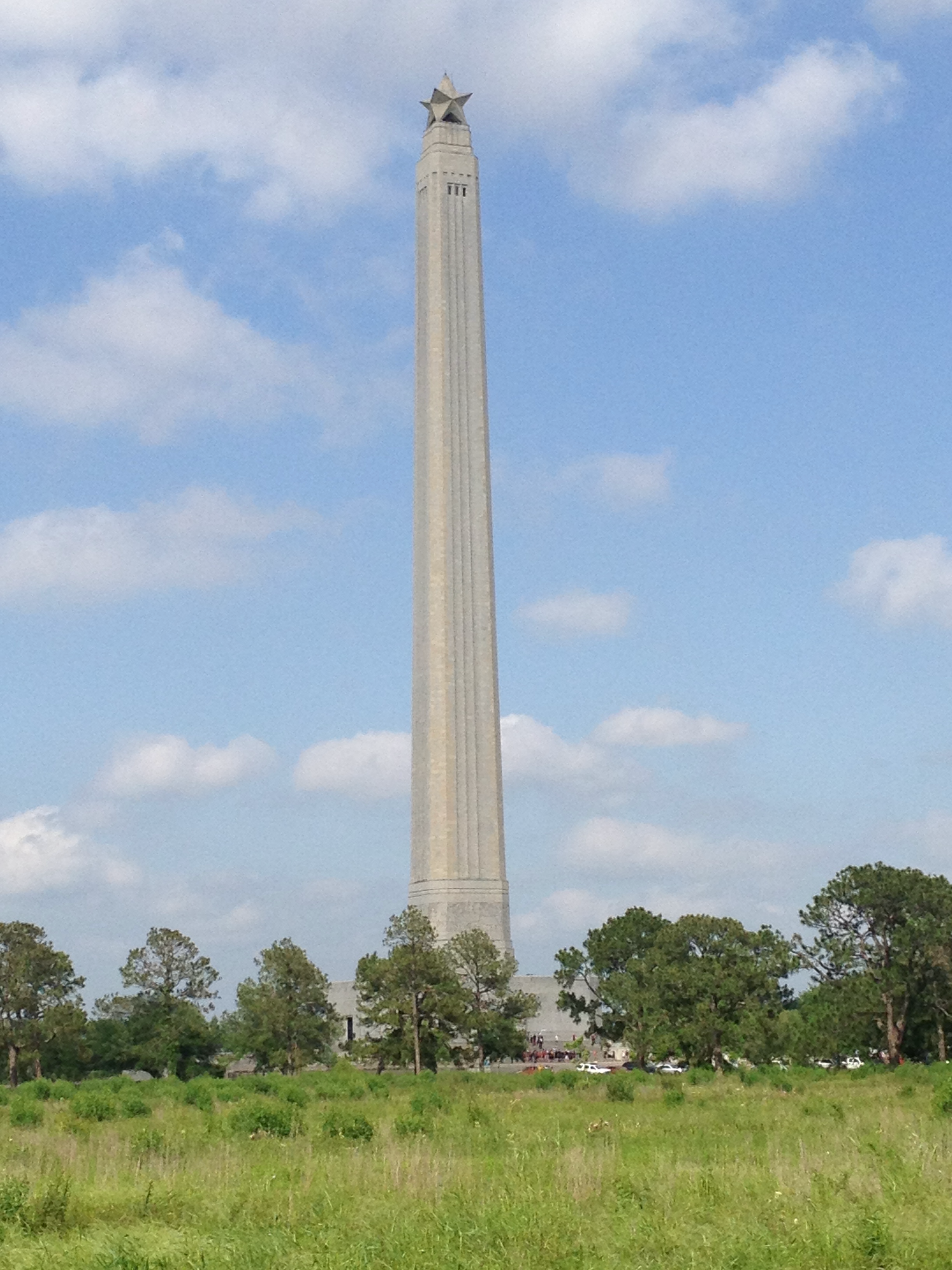 A tall monument with a star at the top, surrounded by greenery and trees, with a parking lot and buildings in the background under a partly cloudy sky.