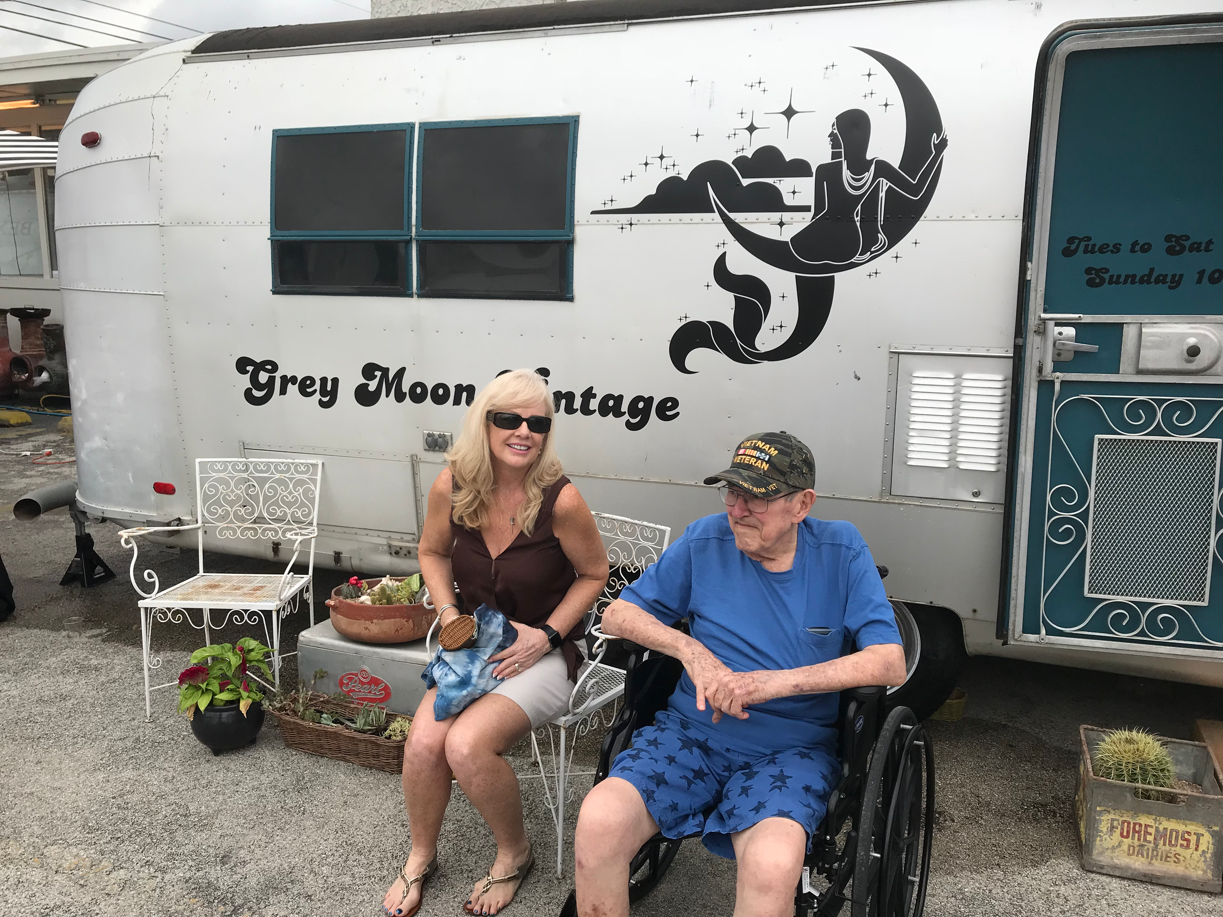 A woman with blonde hair wearing sunglasses and a brown top sitting next to an older man in a wheelchair wearing a blue T-shirt and shorts. They are outdoors in front of a white trailer decorated with a black art design of a mermaid sitting on a crescent moon among stars and clouds. There are outdoor furniture, potted plants, and part of a sign that reads "FOREMOST OATMEAL" visible in the photo.