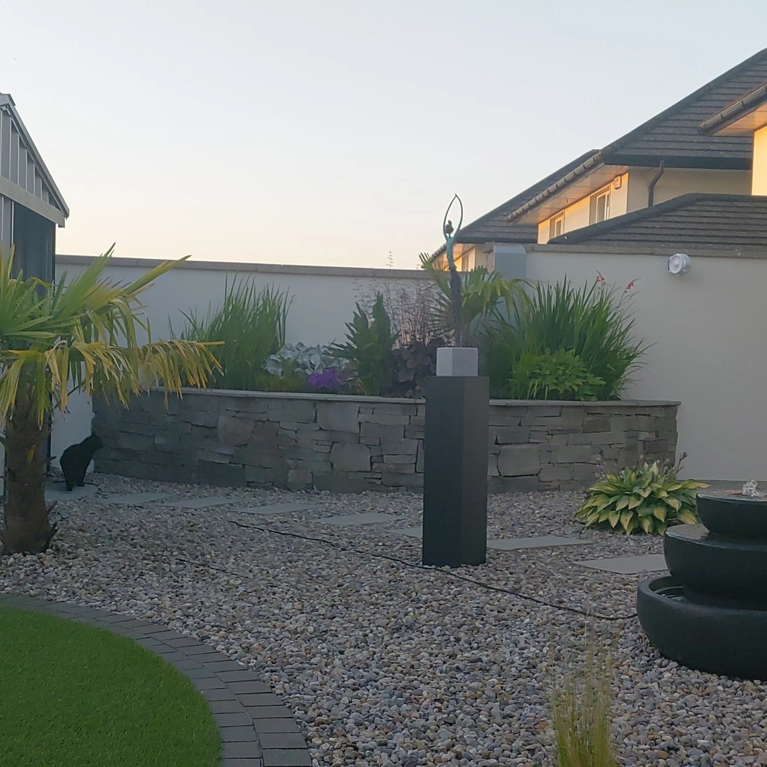 A modern backyard garden with a gravel pathway, green grass, plants, and decorative elements, alongside residential houses.
