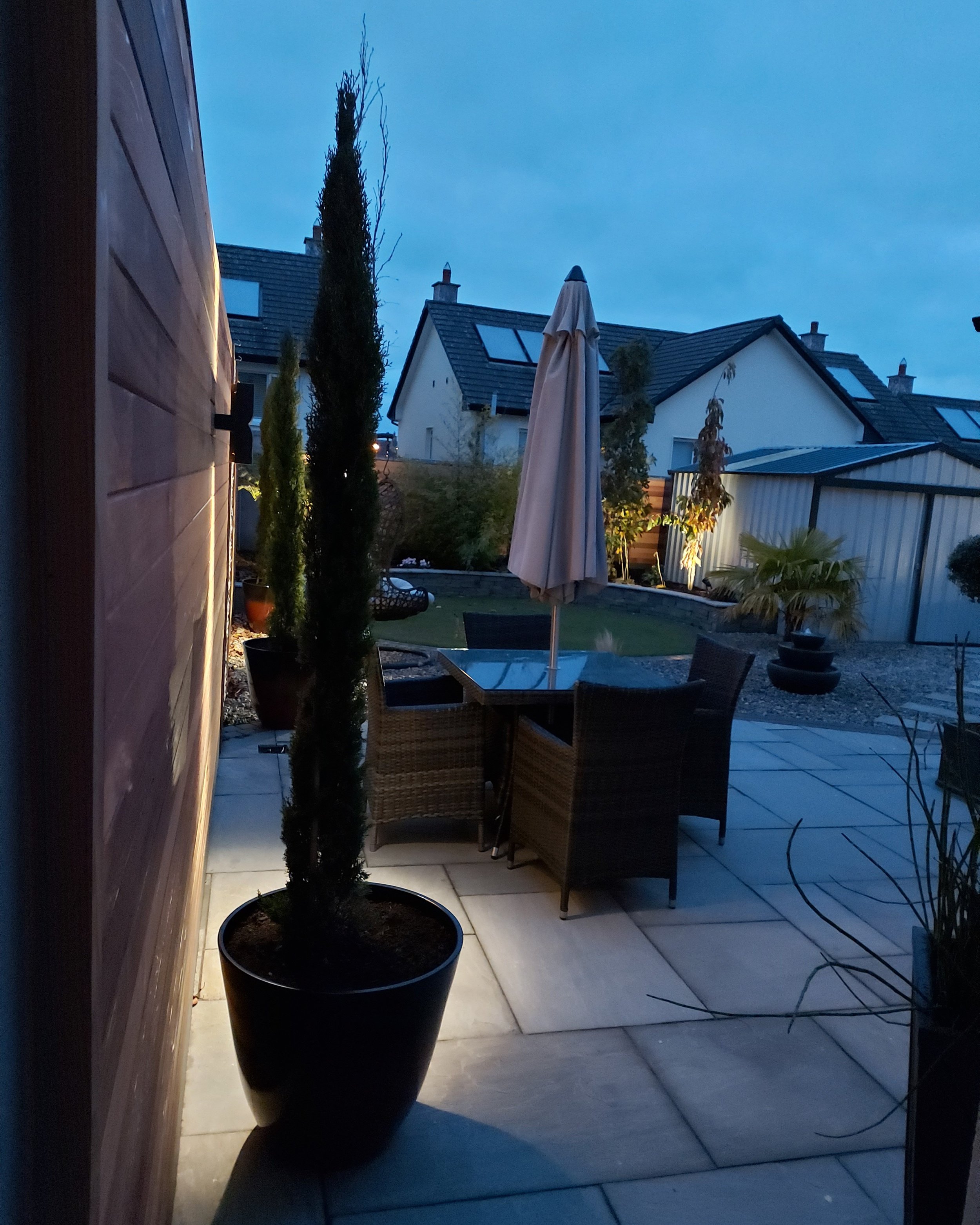 A backyard patio at dusk with potted plants, a dining table with chairs, a closed patio umbrella, and neighboring houses with sloped roofs and skylights.