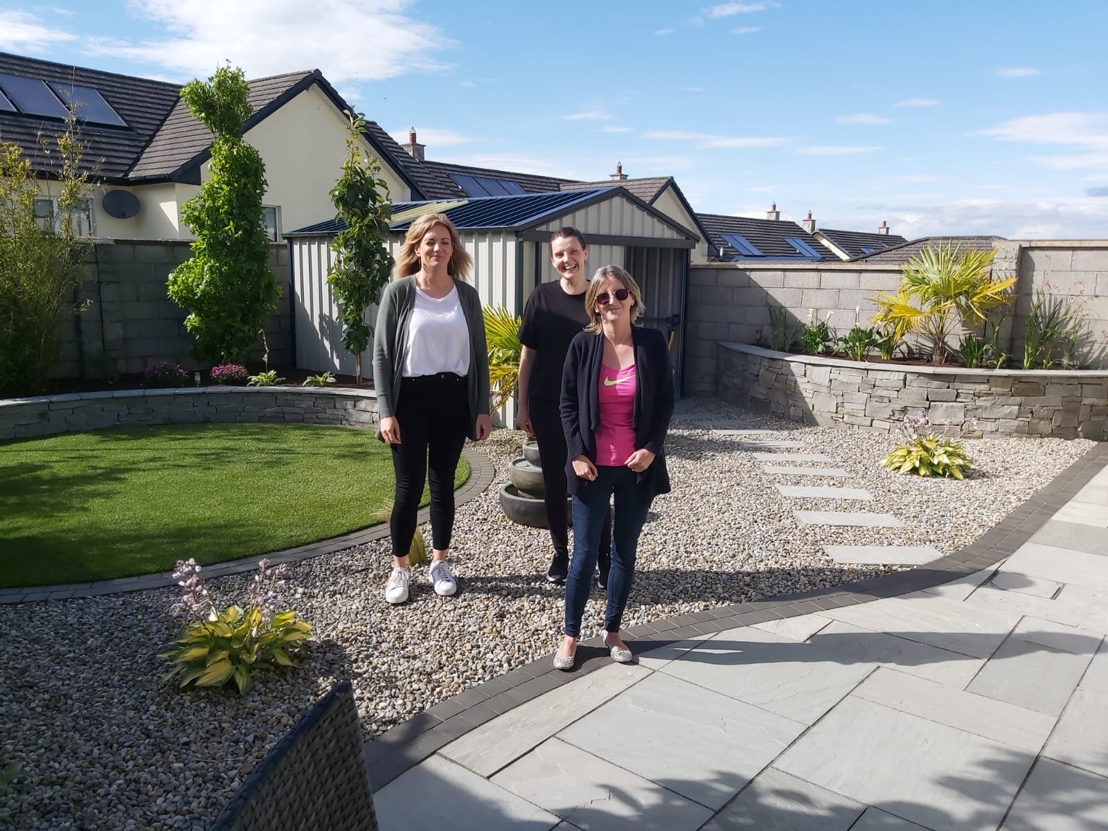 Three women standing in a landscaped backyard garden on a sunny day, with houses and solar panels visible on rooftops in the background.