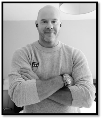 A man with a bald head and beard wearing a light-colored sweater and watch, standing with arms crossed indoors.