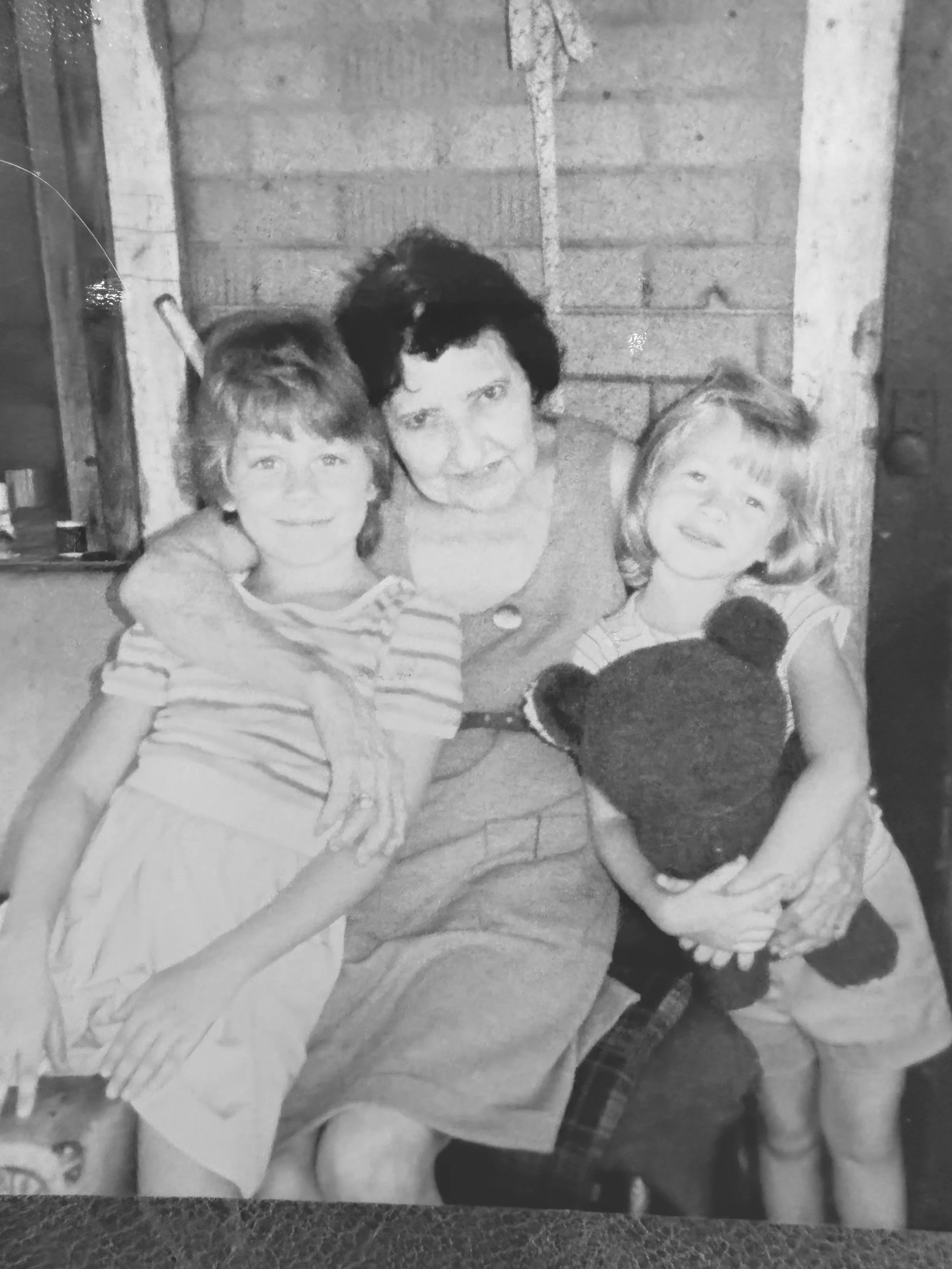 A woman with short dark hair and light skin sitting on the floor with two young child, all smiling at the camera. The children have light skin. The girl has light-colored hair and is wearing a striped shirt. They are in a room with a brick wall in the background.