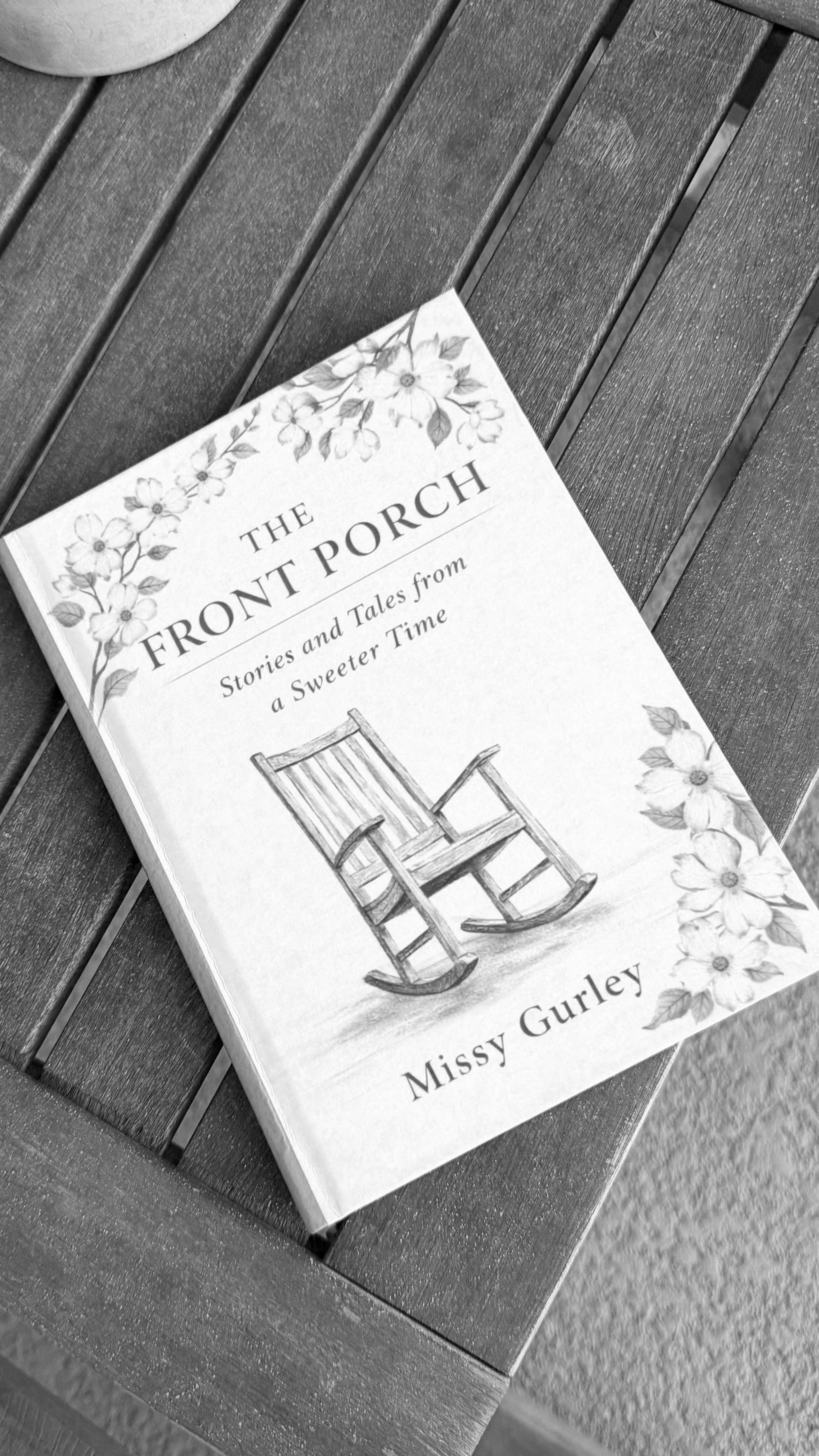 A book titled 'The Front Porch: Stories and Tales from a Sweeter Time' by Missy Gurley resting on a wooden bench.