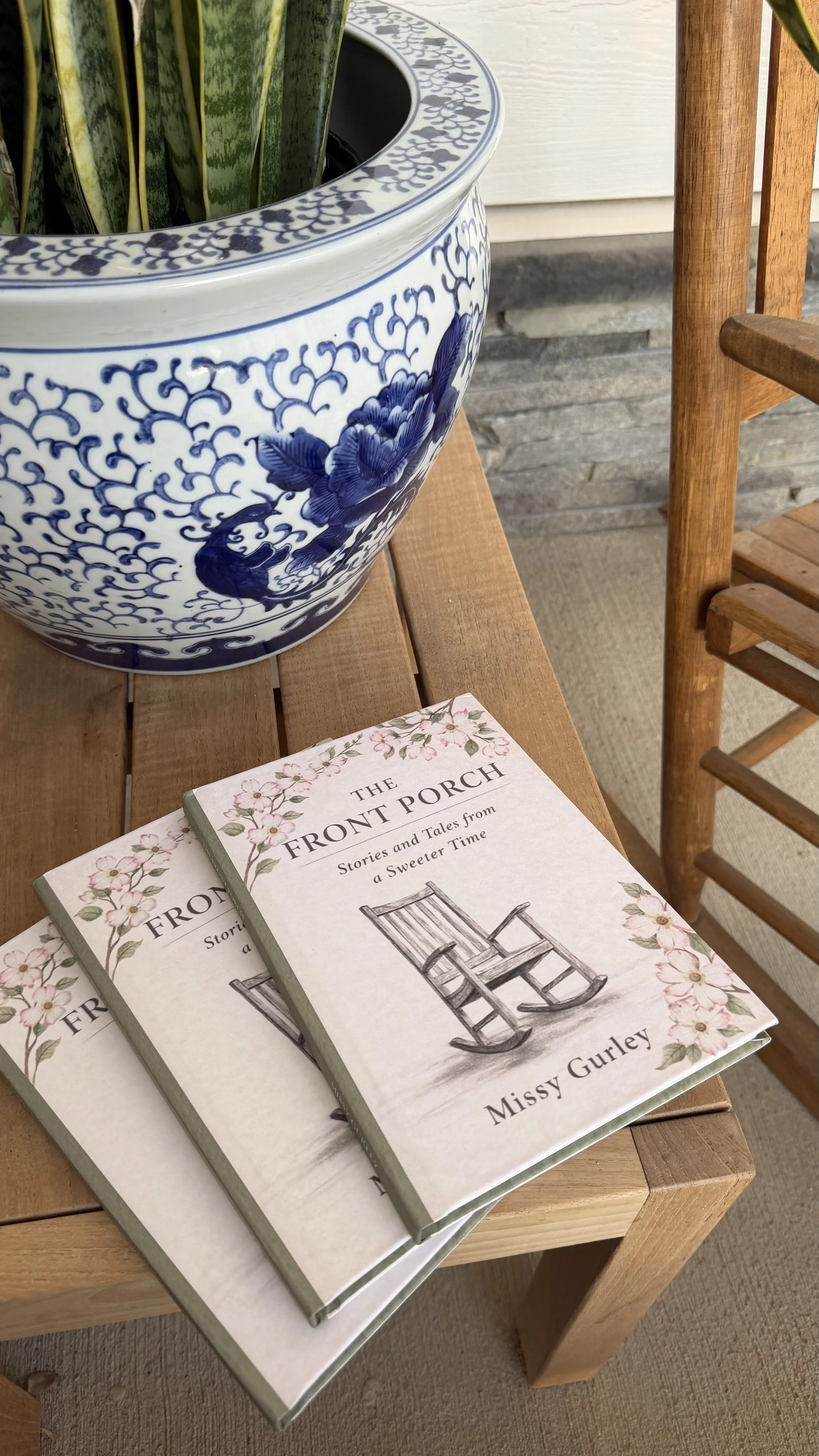 A large porcelain blue and white planter with a chinoiserie design, resting on a wooden table. The table also holds a stack of books titled 'The Front Porch' by Missy Gurley. Part of a wooden chair and a stone wall background are visible.