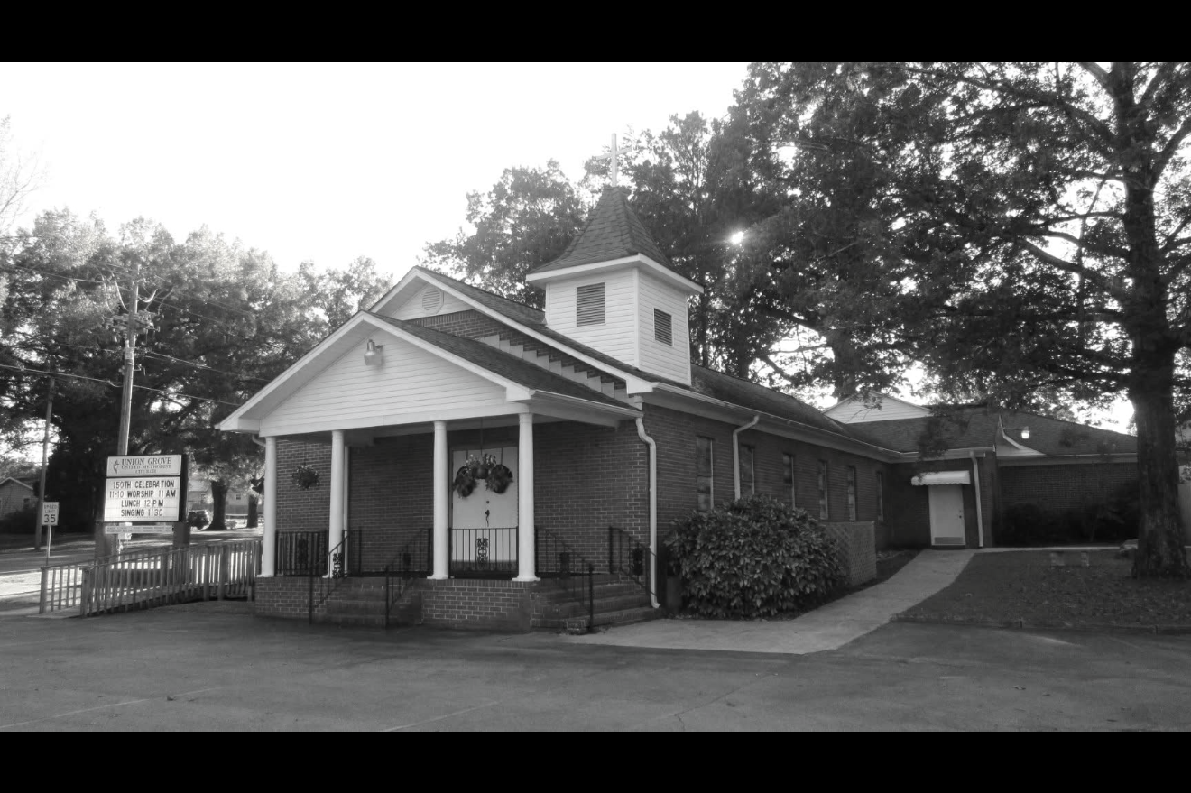 Black and white photo of a church with a brick facade, a small front porch with white columns, steps leading up to it, and a small steeple on the roof surrounded by trees. Southern Alabama church