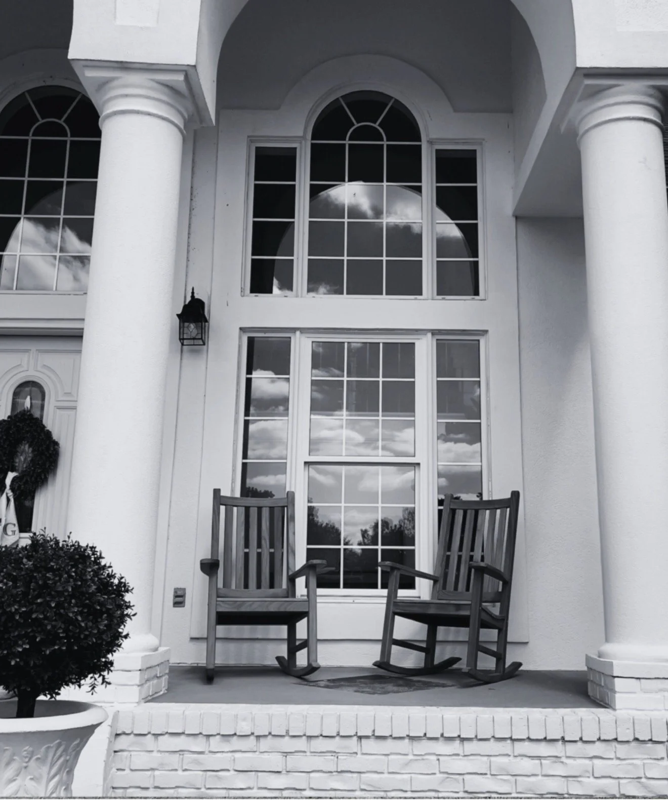 Black and white photo of front porch of a house with two rocking chairs, large window reflecting clouds, white columns, and decorative urn plant at the corner.