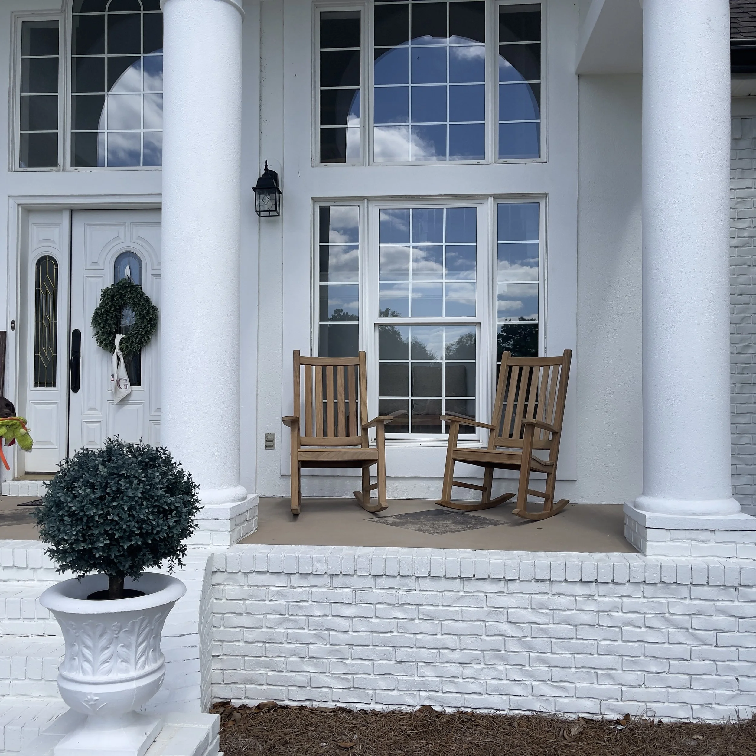 Front of a white house with large windows, white brick porch, a front door with decorative glass, a wreath, a black lantern, potted bush, and wooden chairs outside.
