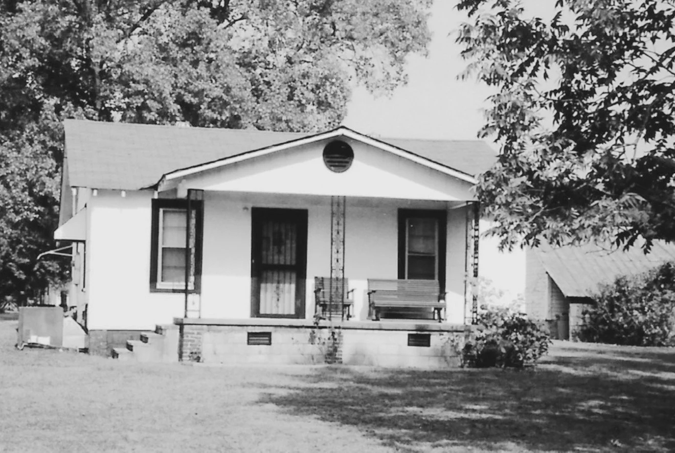 Black and white photo of a small house with a front porch, two open windows, a door, and a pitched roof, surrounded by trees and grassy yard.