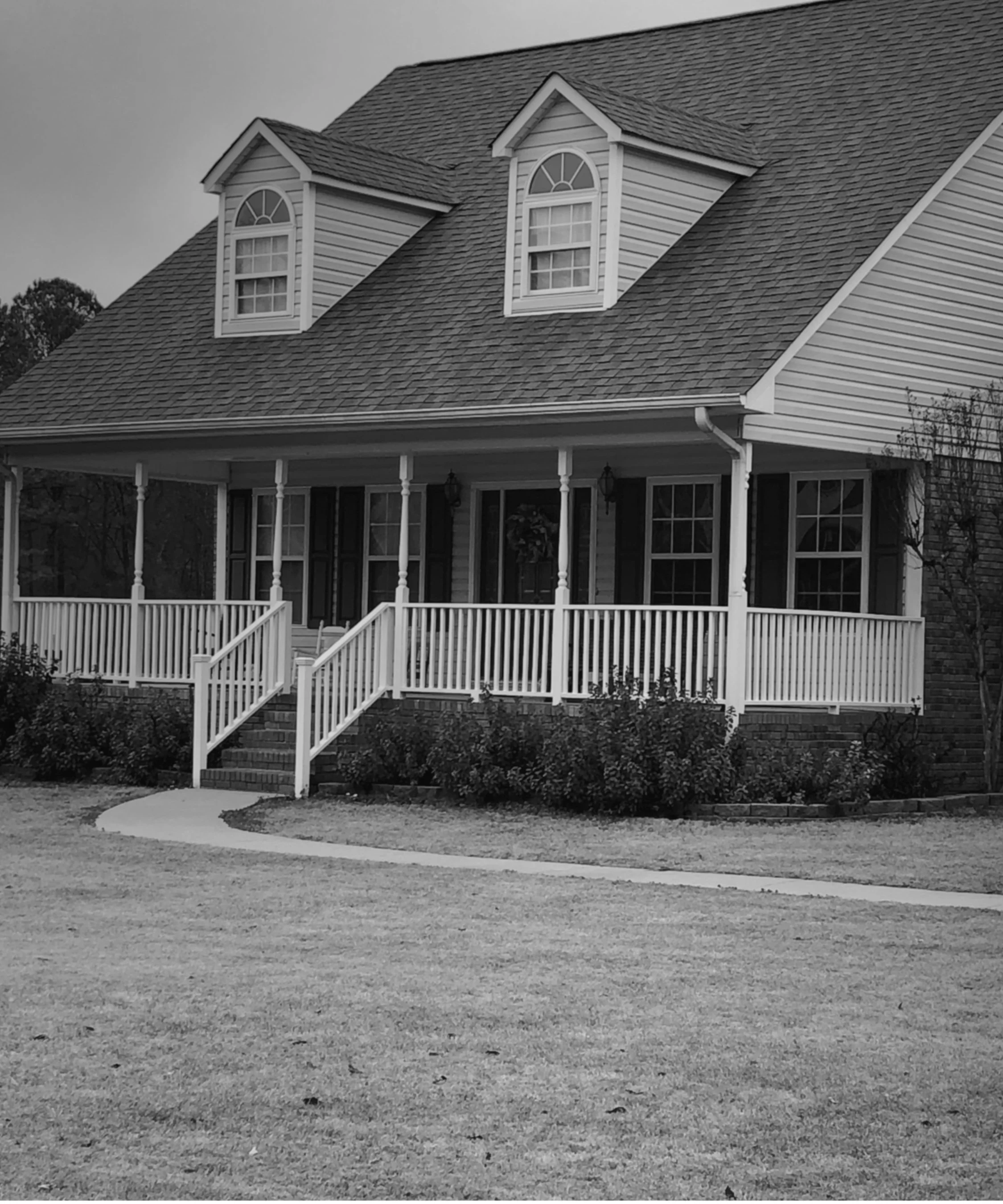 A house with a gabled roof, dormer windows, a porch with white railings and columns, and a front yard with grass and bushes.