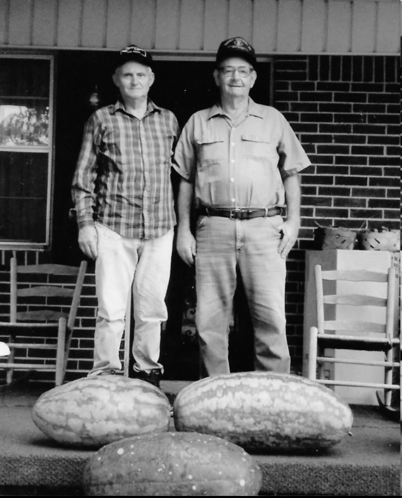 Two older men standing behind large watermelons on the ground, with chairs and a brick wall in the background, in a black-and-white photo.
