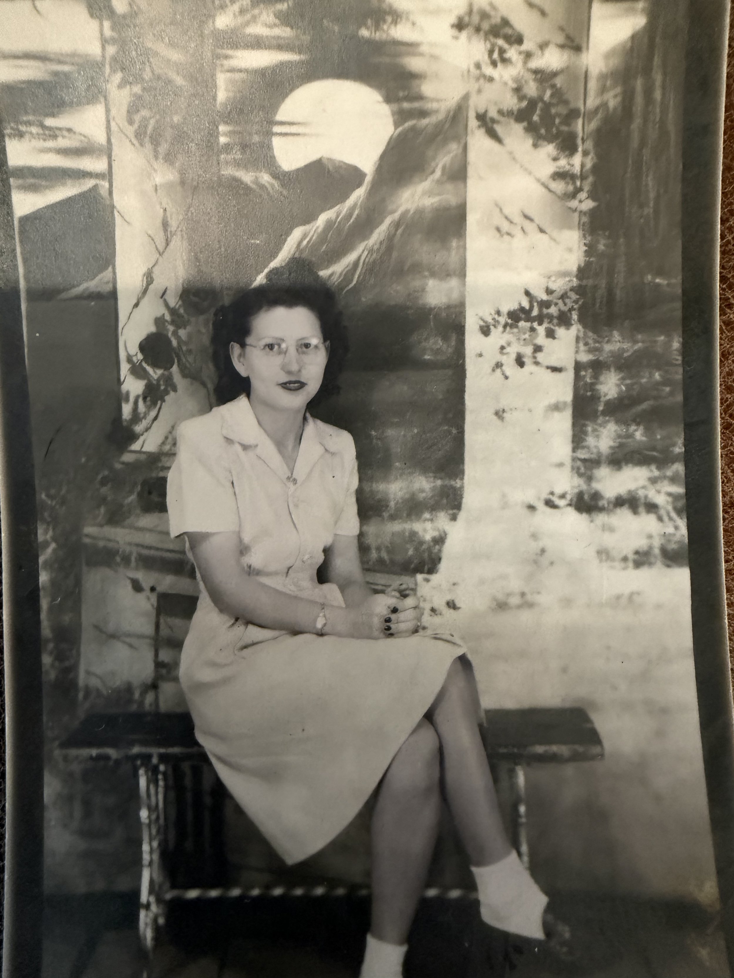 Black and white photo of a woman sitting on a chair outdoors with a mountain landscape and a setting sun in the background. She is wearing glasses, a light-colored dress, and dark lipstick, with her hands clasped on her lap.