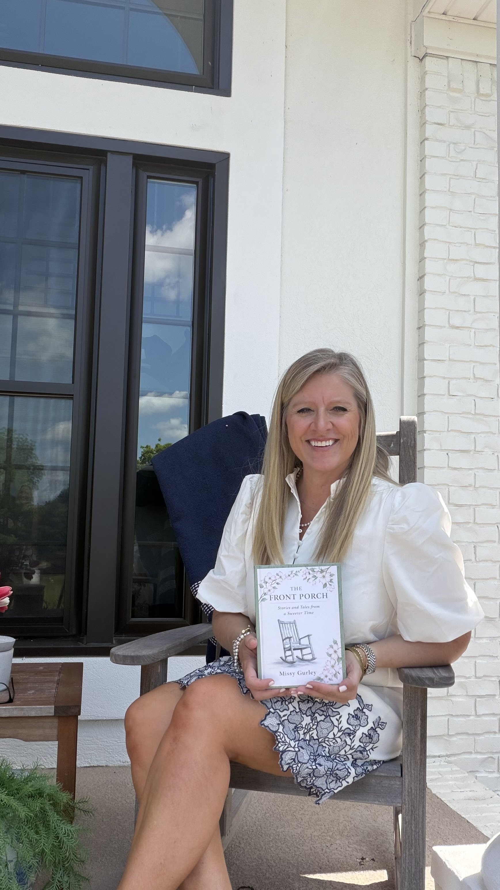 Author, Missy Gurley,  sitting on a rocking chair outdoors, holding her book titled "The Front Porch" by Missy Gurley, with a white house and large windows in the background.