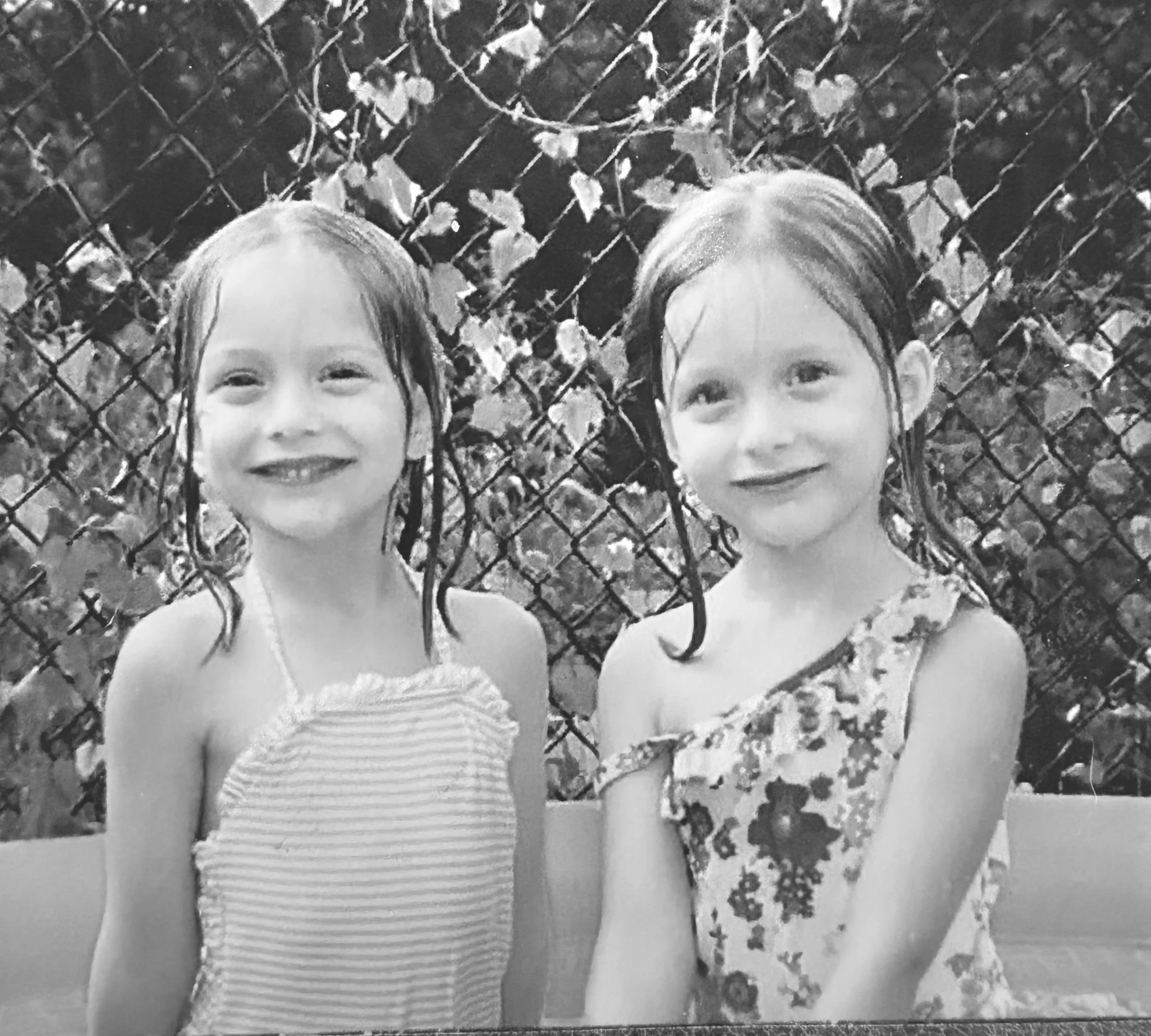 Two young girls with long hair and light skin are smiling while standing in front of a chain-link fence that has some ivy and leaves growing on it. One girl is wearing a striped tank top, and the other girl is wearing a light-colored tank top.