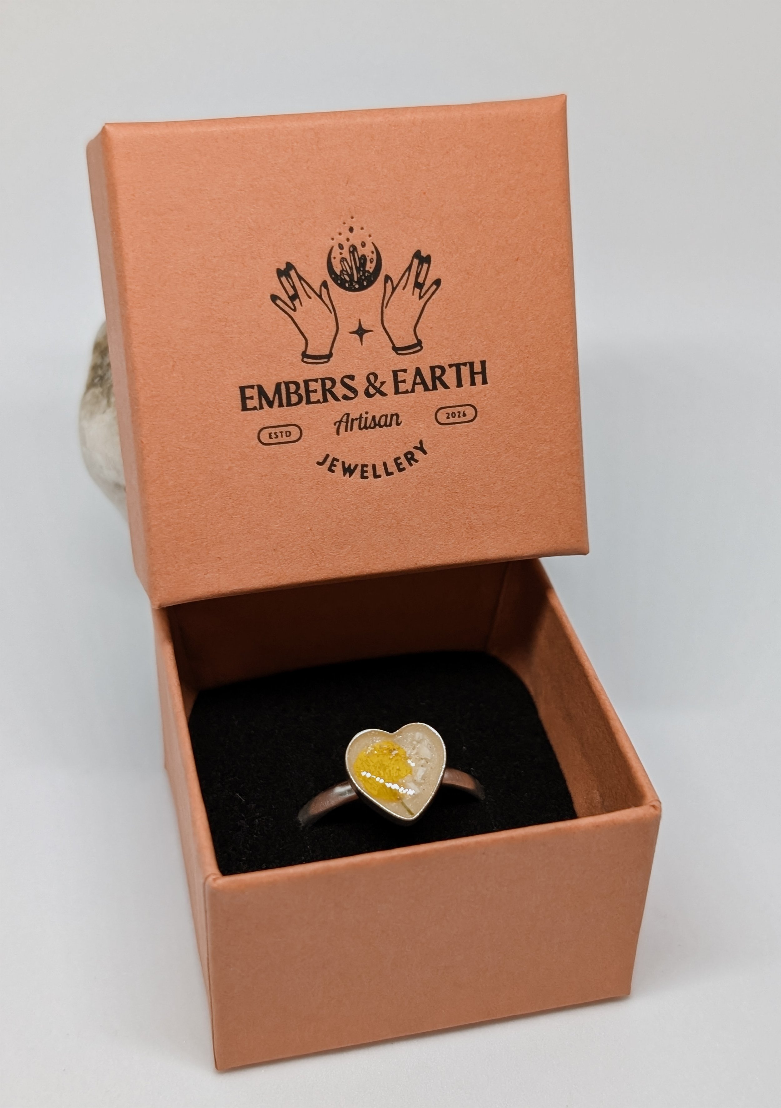 A ring with a heart-shaped face inside a pink jewelry box. The face has yellow and white details. The jewelry box has a logo on the inside lid with two hands, a crescent moon, and the words "Embers & Earth," "Artisan Jewelry," and "ESTD 2016."