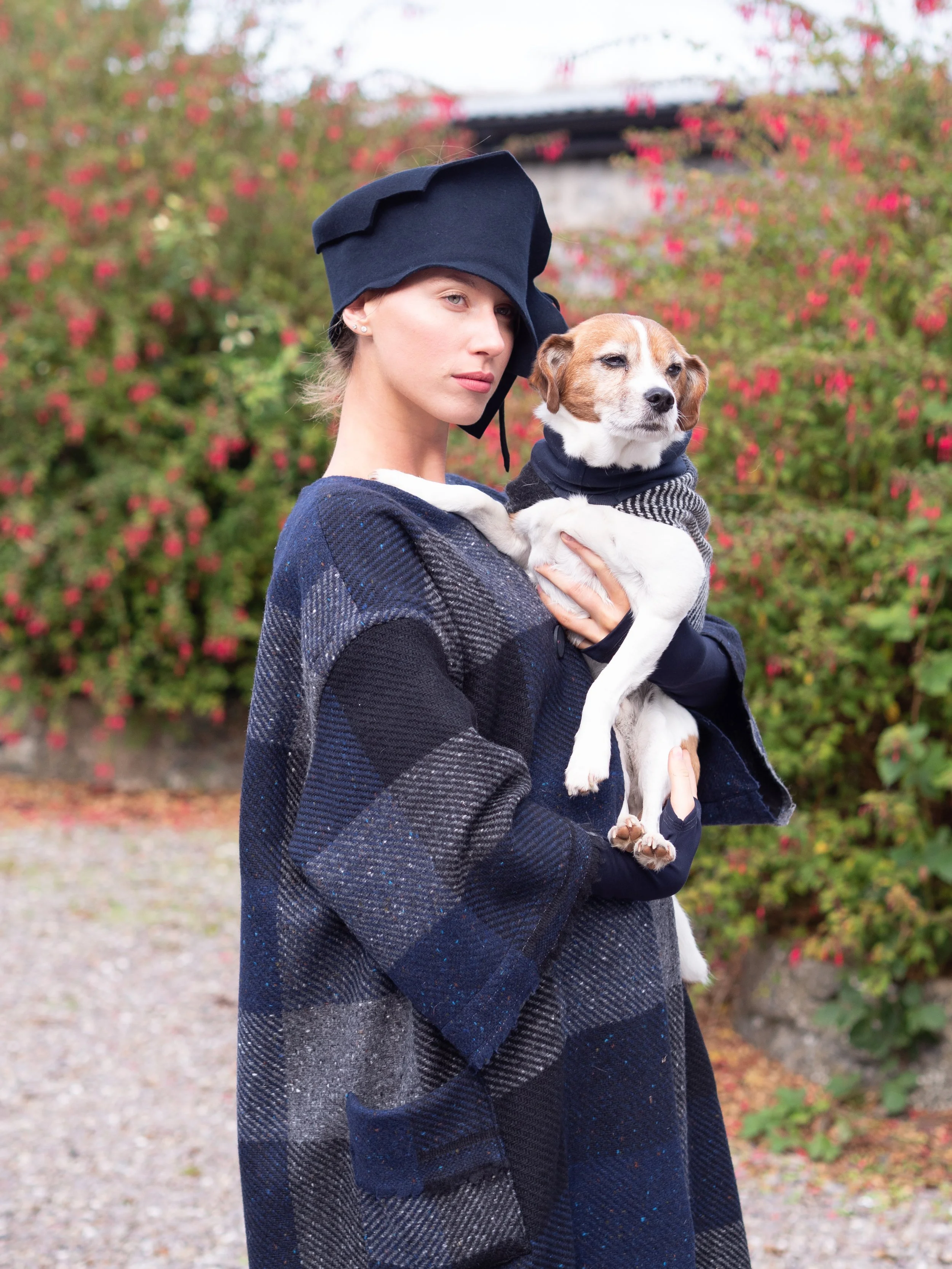 A woman in a plaid coat and a nurse cap holding a dog with a black scarf in front of a colorful bush with pink and red flowers. Mise Tusa Clarinbridge design galway ireland meritta