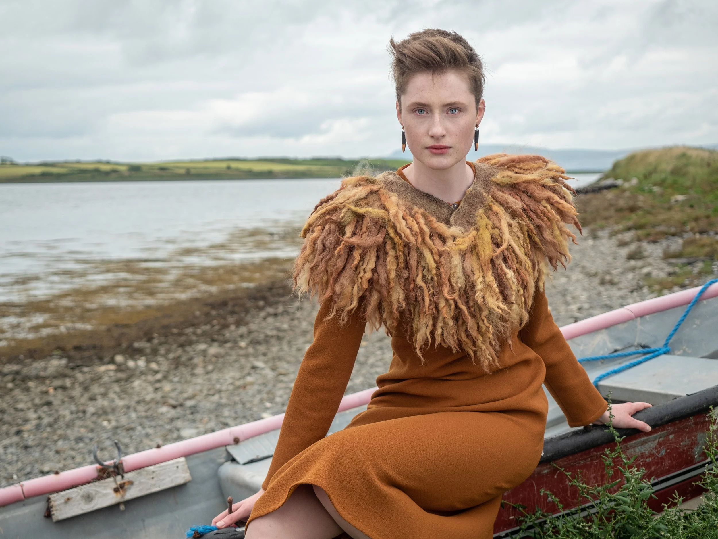 A young woman with short, styled hair and blue eyes sitting on a boat near a rocky shoreline, wearing a textured, multicolored sweater and brown pants, Mise Tusa Clarinbridge design galway ireland meritta