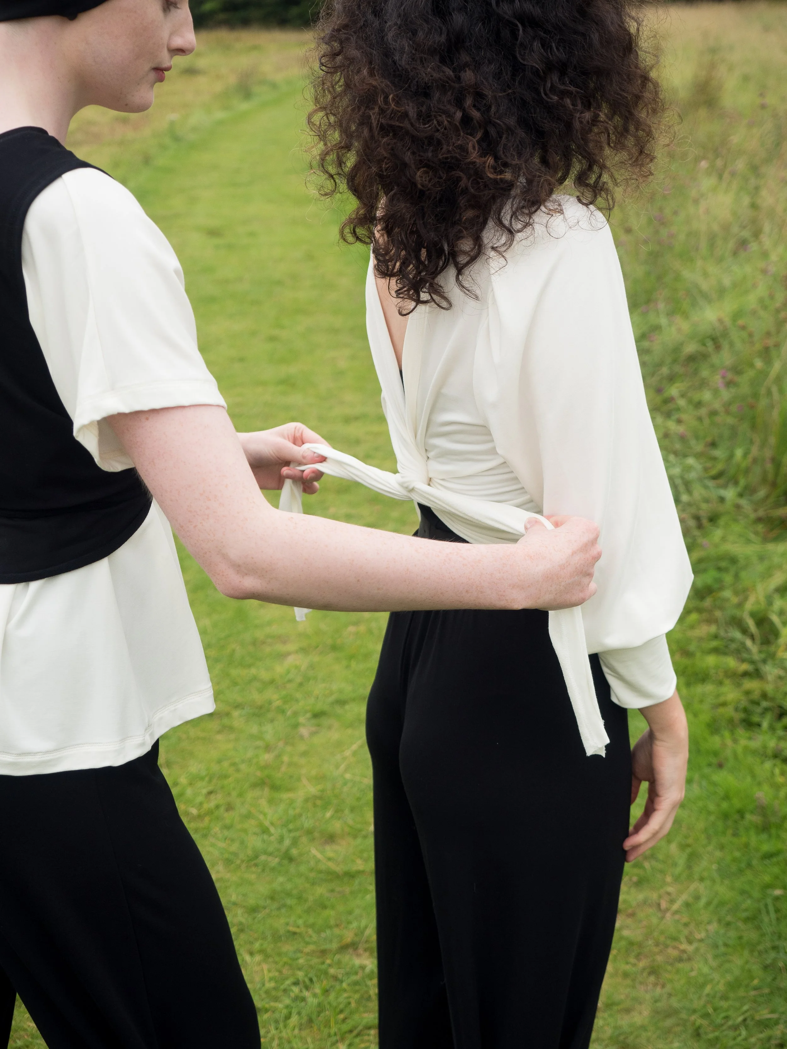 Two women standing outdoors on a grassy area, holding hands, with one woman tying the other's shirt at the waist. Mise Tusa Clarinbridge design galway ireland meritta