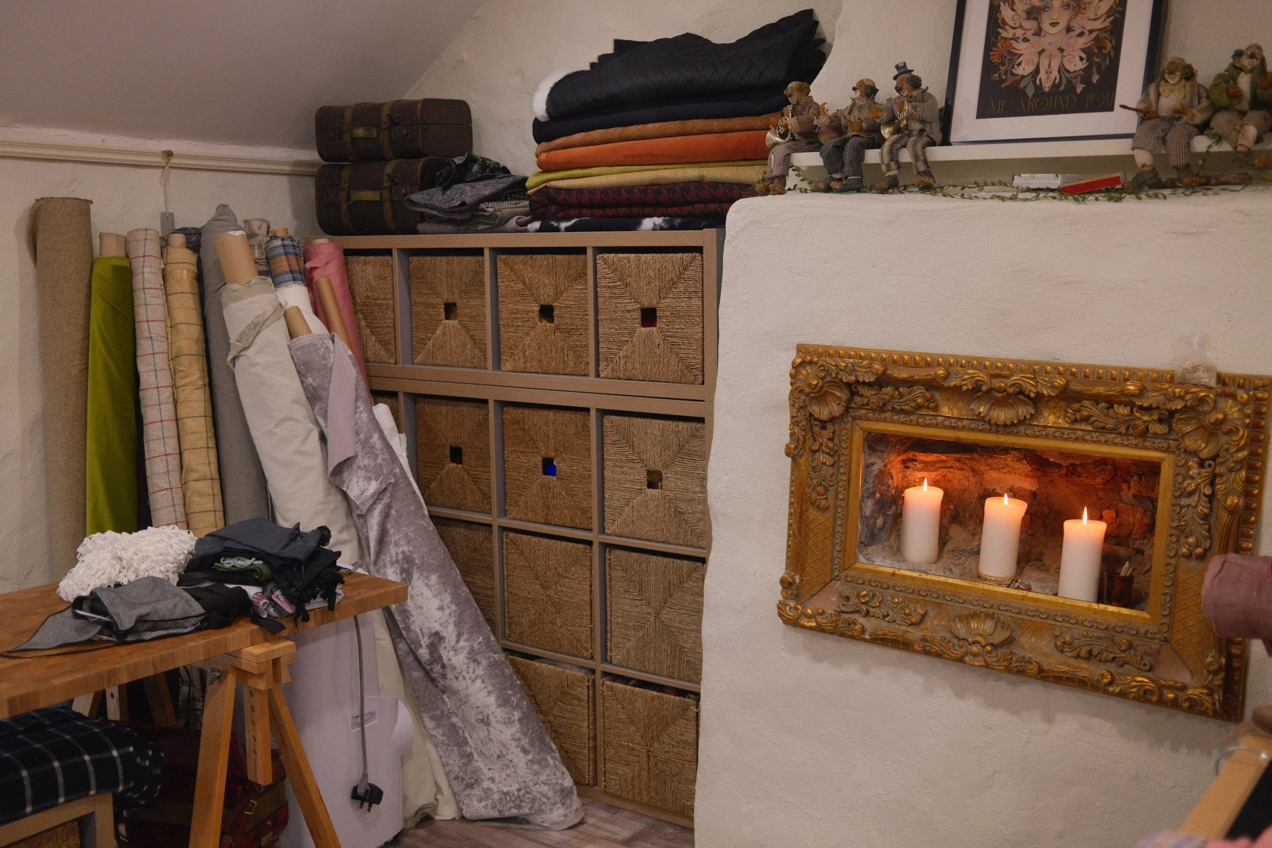 A cluttered room with a wooden table filled with clothing, fabric rolls, and an iron. Above the table, a storage shelf holds folded blankets and cushions. To the Mise Tusa Clarinbridge design galway ireland meritta