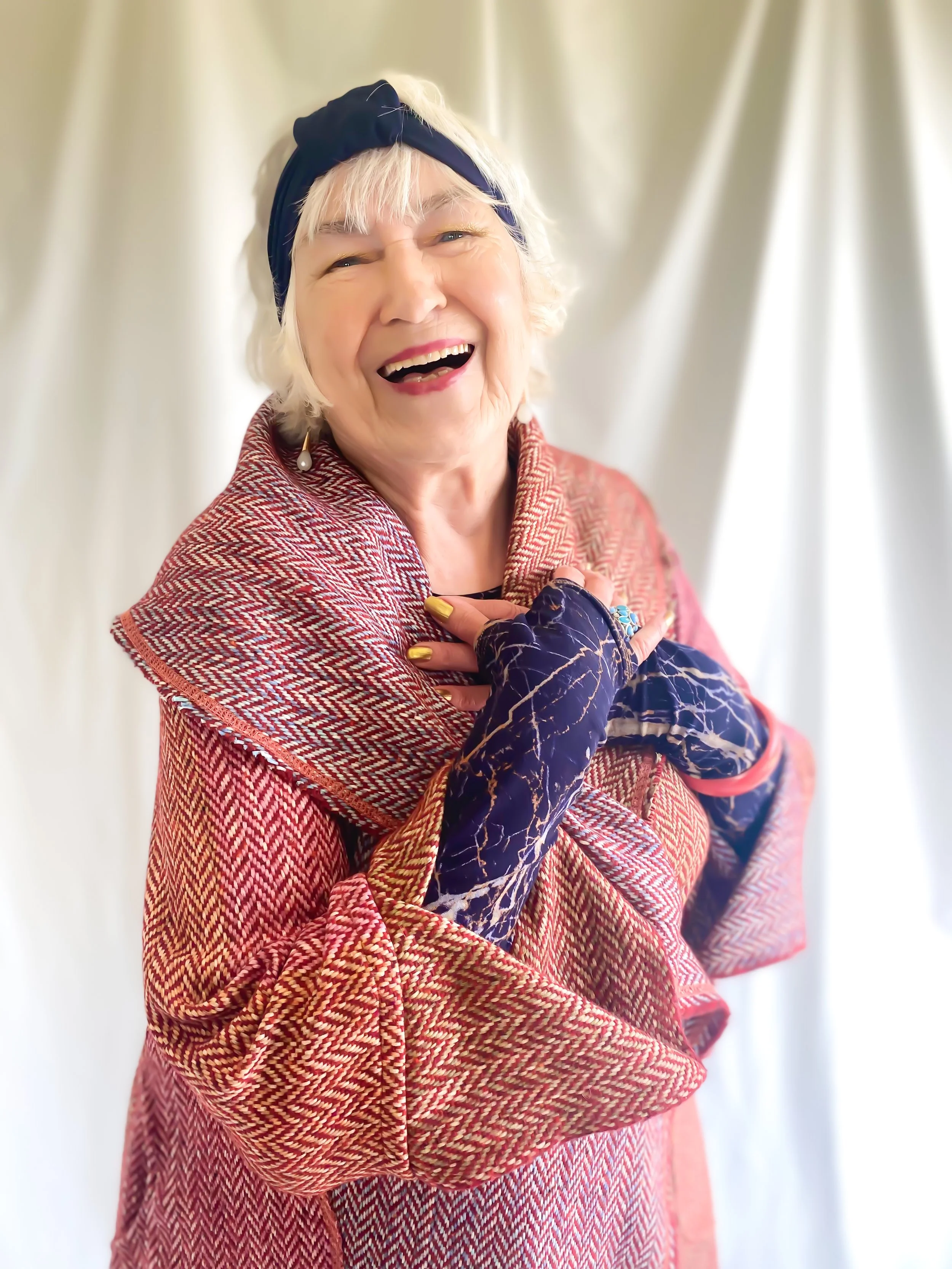 An elderly woman smiling and laughing, wearing a blue headband, earrings, a red patterned shawl, and colorful gloves, standing in front of a light-colored curtain. Mise Tusa Clarinbridge design galway ireland meritta