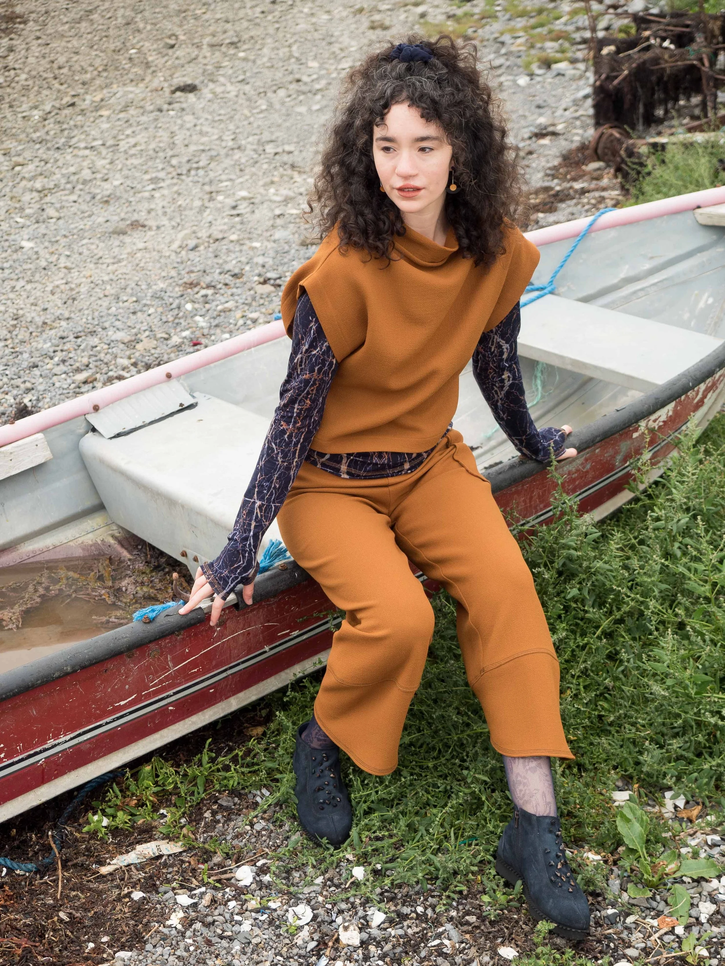 A woman with curly hair, wearing a brown outfit with patterned sleeves, sitting on the edge of a small boat on a rocky shoreline with green plants and old rusted metal debris in the background. Mise Tusa Clarinbridge design galway ireland meritta