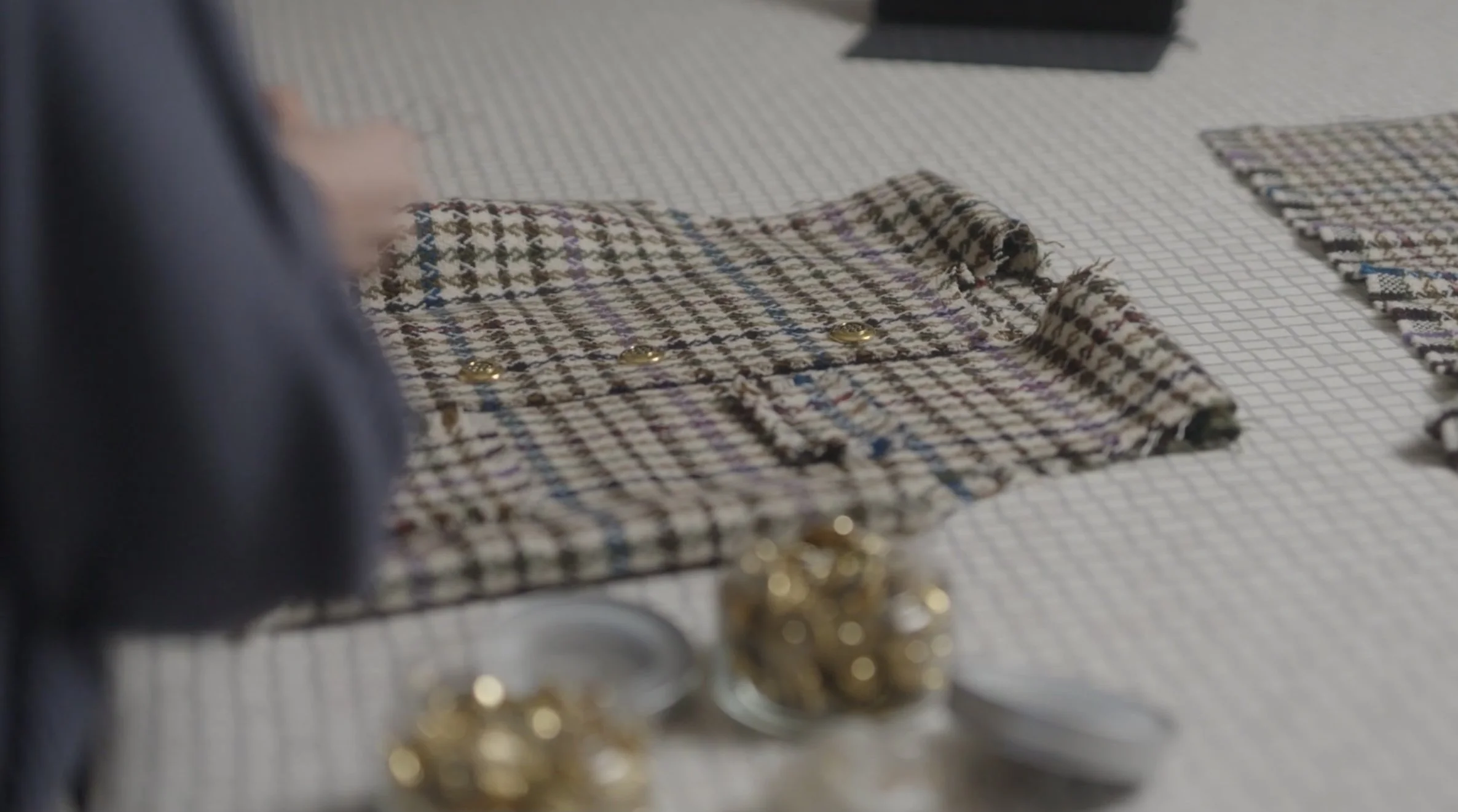 Close-up of a fabric piece with small gold buttons on a table with a grid pattern, with additional fabric pieces nearby and blurred gold objects in the foreground. Mise Tusa Clarinbridge design galway ireland meritta