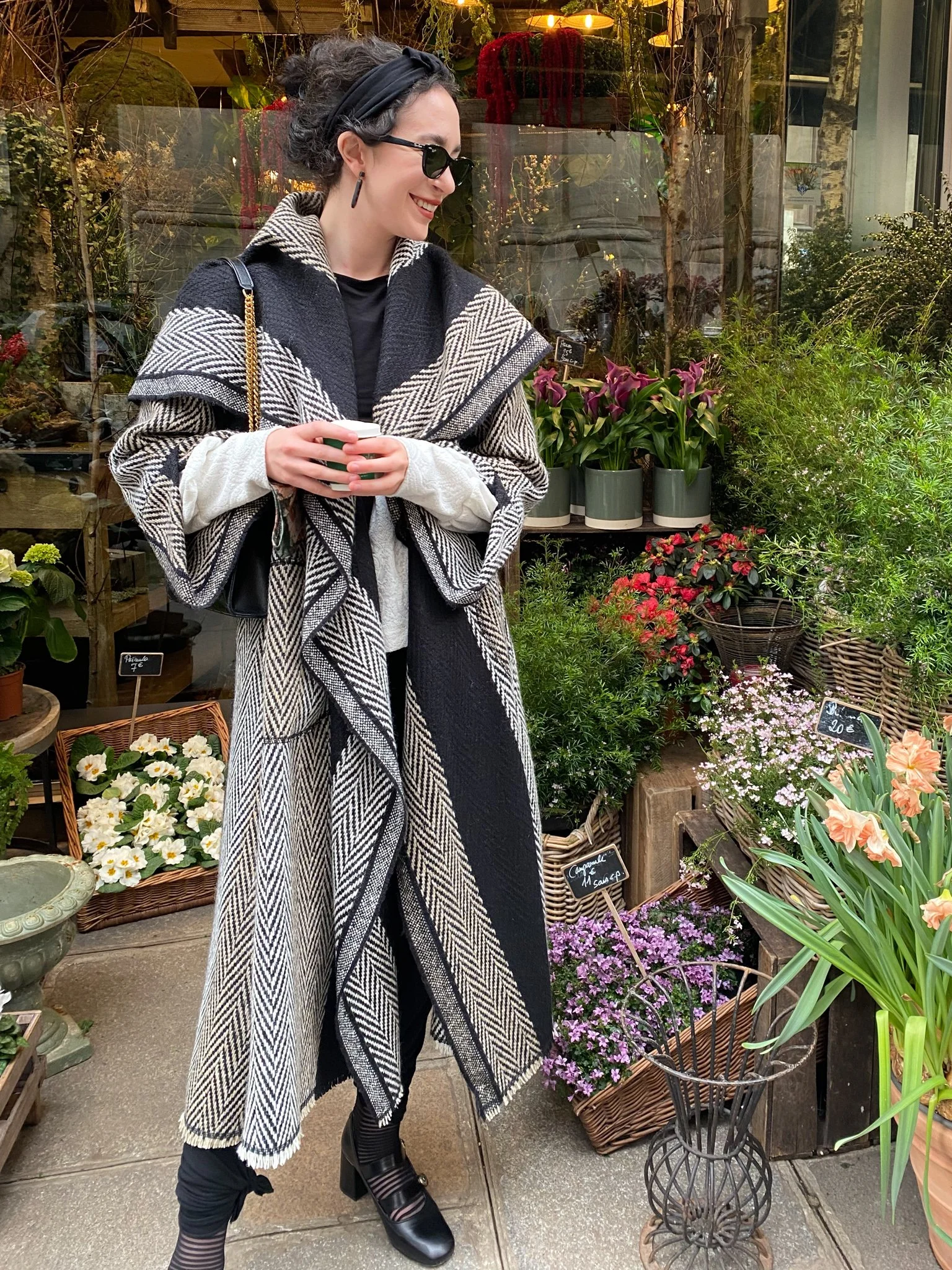 A woman with dark curly hair, wearing a black headband, sunglasses, and a patterned black and white coat, is standing in a flower shop, smiling and holding Mise Tusa Clarinbridge design galway ireland meritta