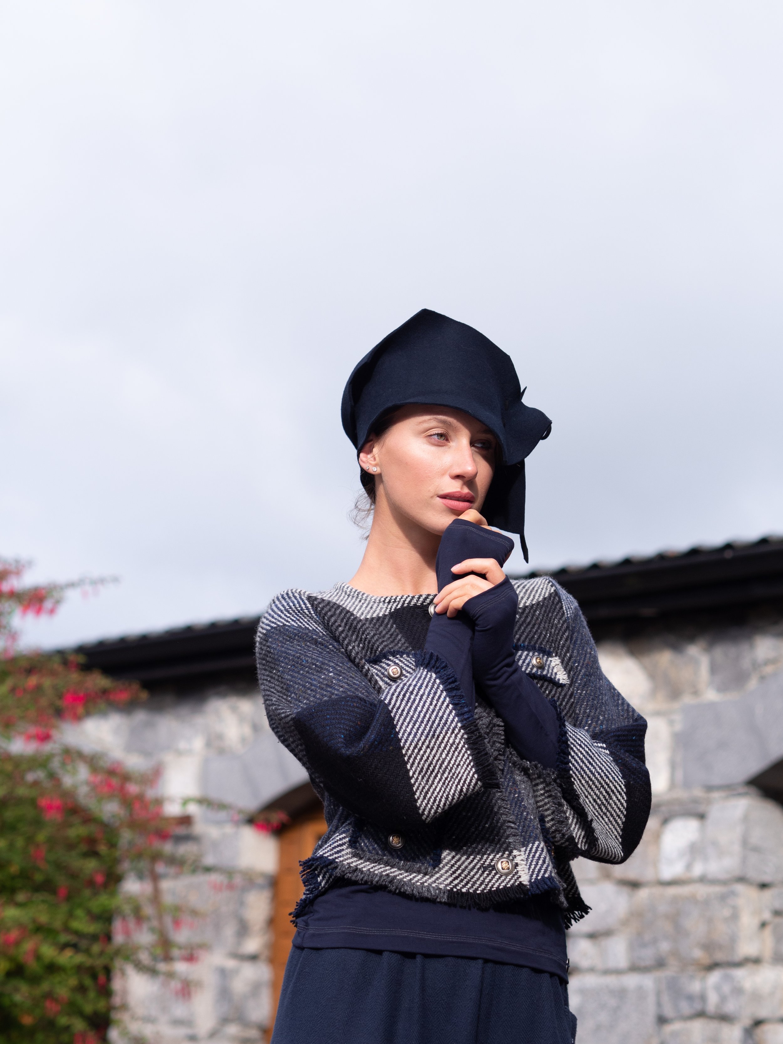  Woman wearing a dark hat, gray plaid coat, and dark gloves, standing outdoors with a stone wall and flowers in the background. Mise Tusa Clarinbridge design galway ireland meritta