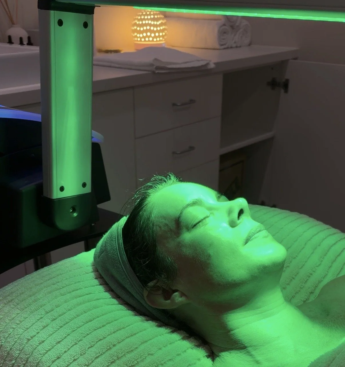 A woman lying on a treatment bed with her eyes closed, undergoing an LED light therapy session with a green light directed at her face in a clinical setting.