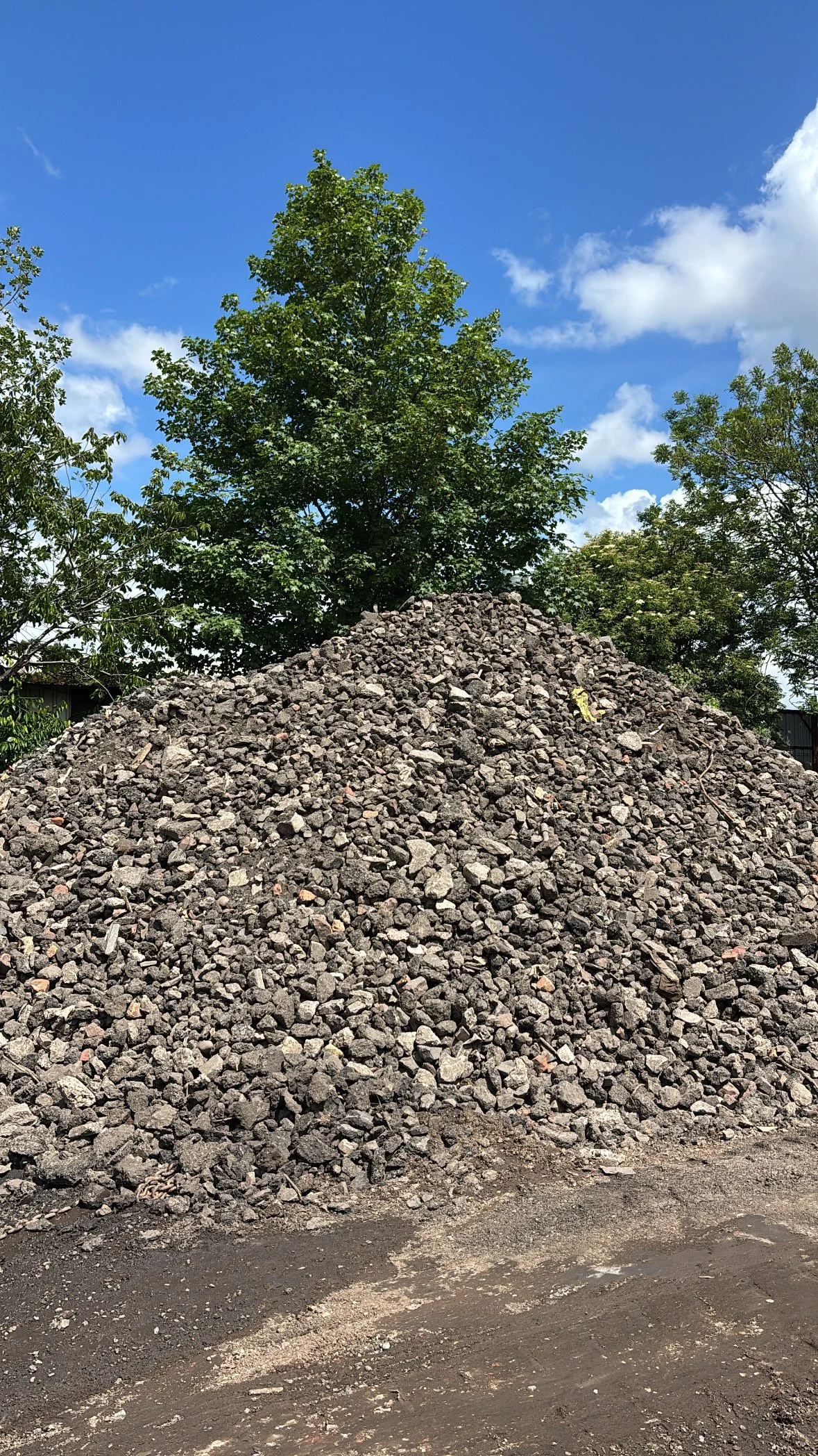 A pile of gravel or small rocks in front of a green tree under a blue sky with clouds.