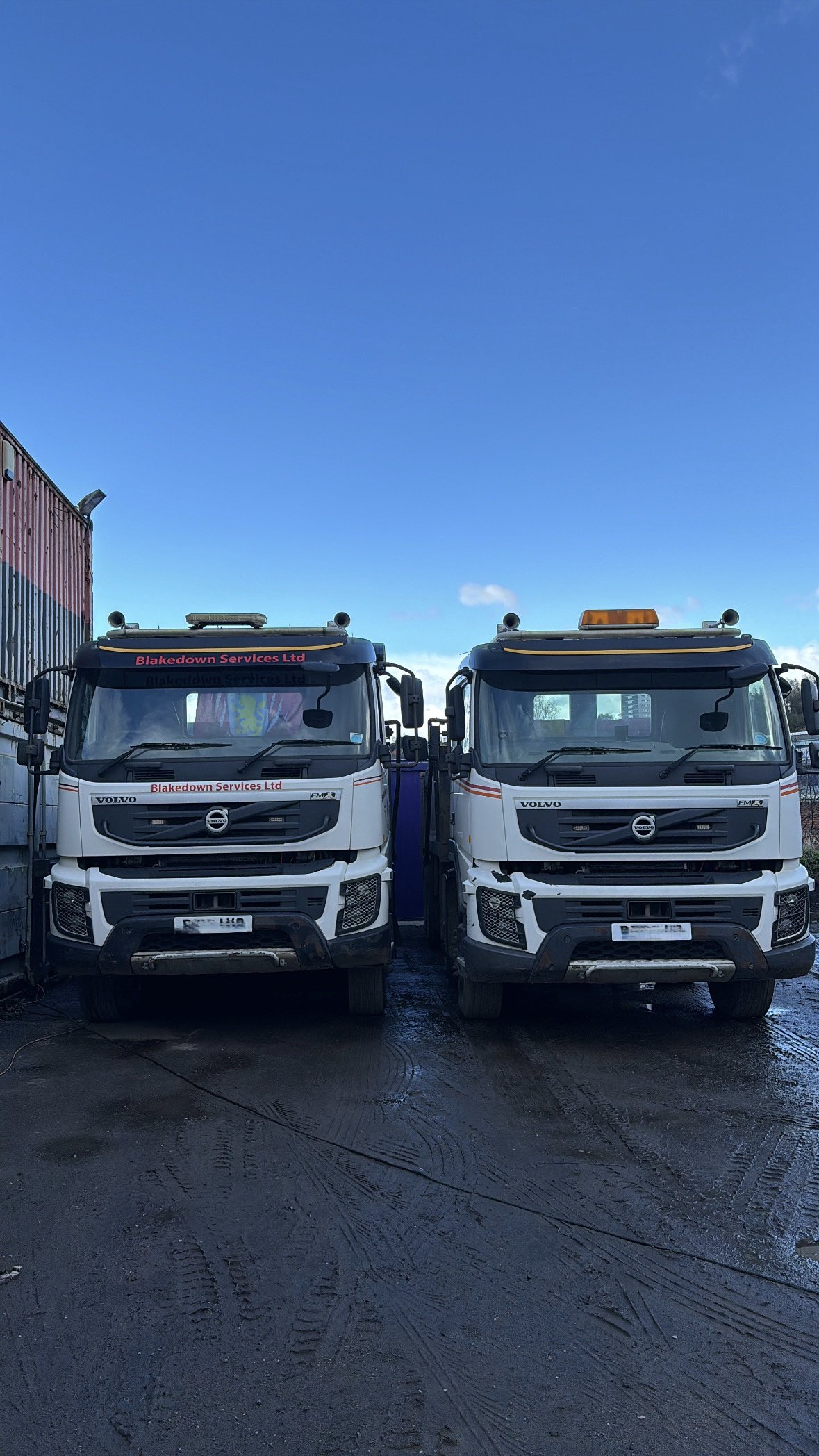 Two white Volvo trucks parked side by side in an outdoor lot, with clear blue sky overhead.