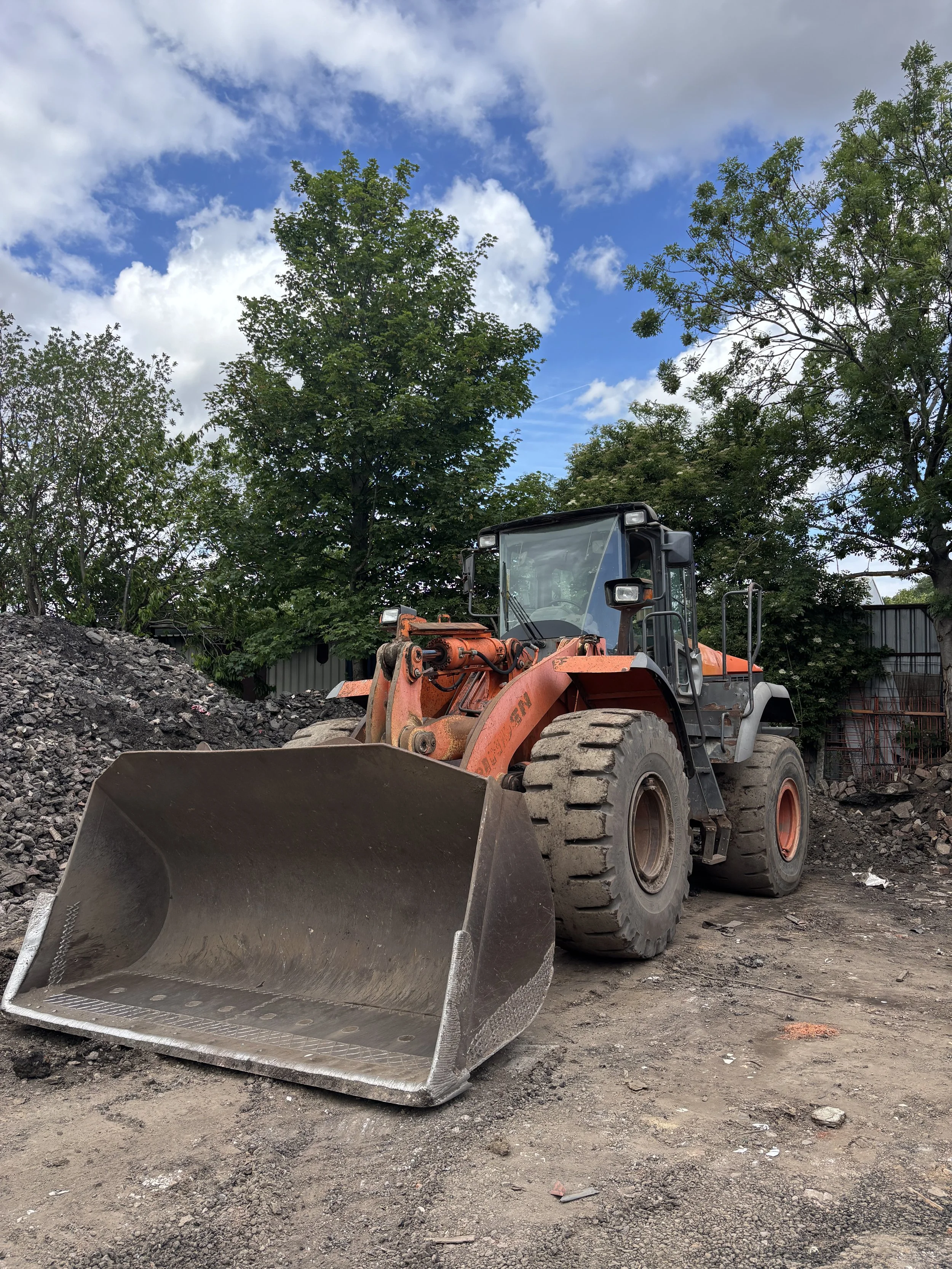 Construction site with an orange front loader digging into dirt, green trees in the background, partly cloudy sky.